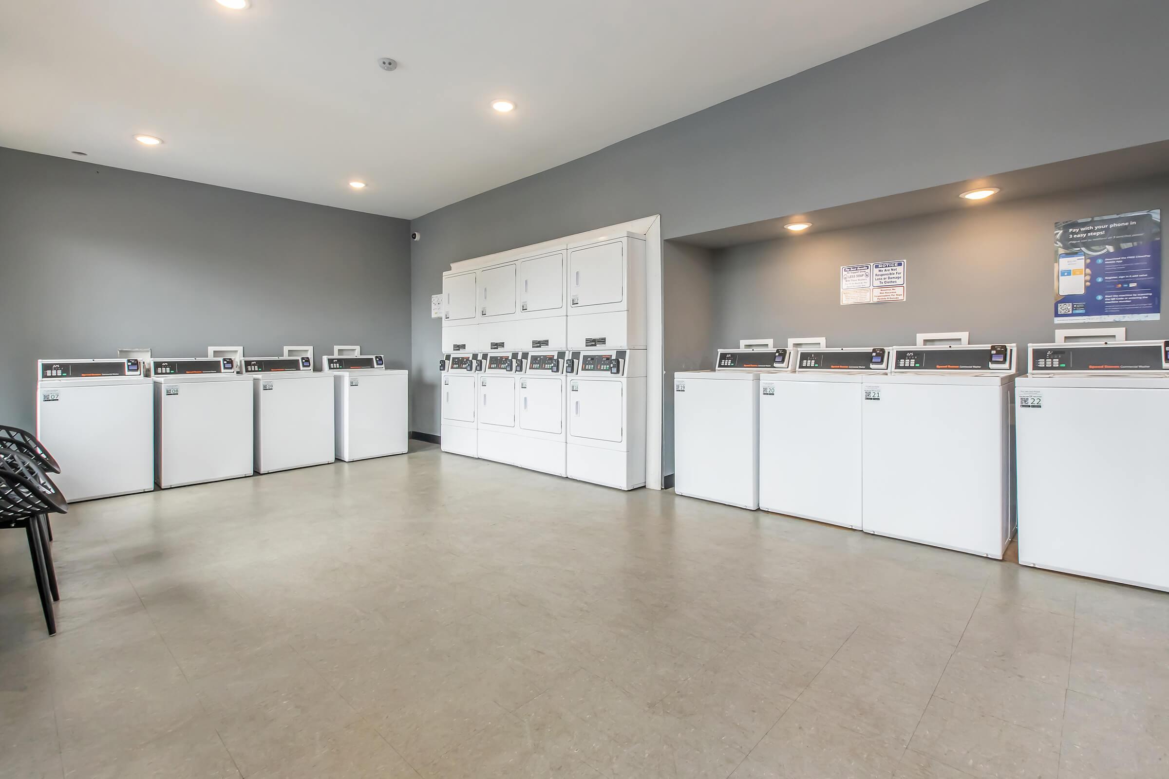 A clean and modern laundromat featuring multiple white washing machines and dryers spaced along the walls. There are chairs for sitting, with gray walls and bright lighting creating a welcoming atmosphere. A bank of machines is positioned opposite a white storage unit.