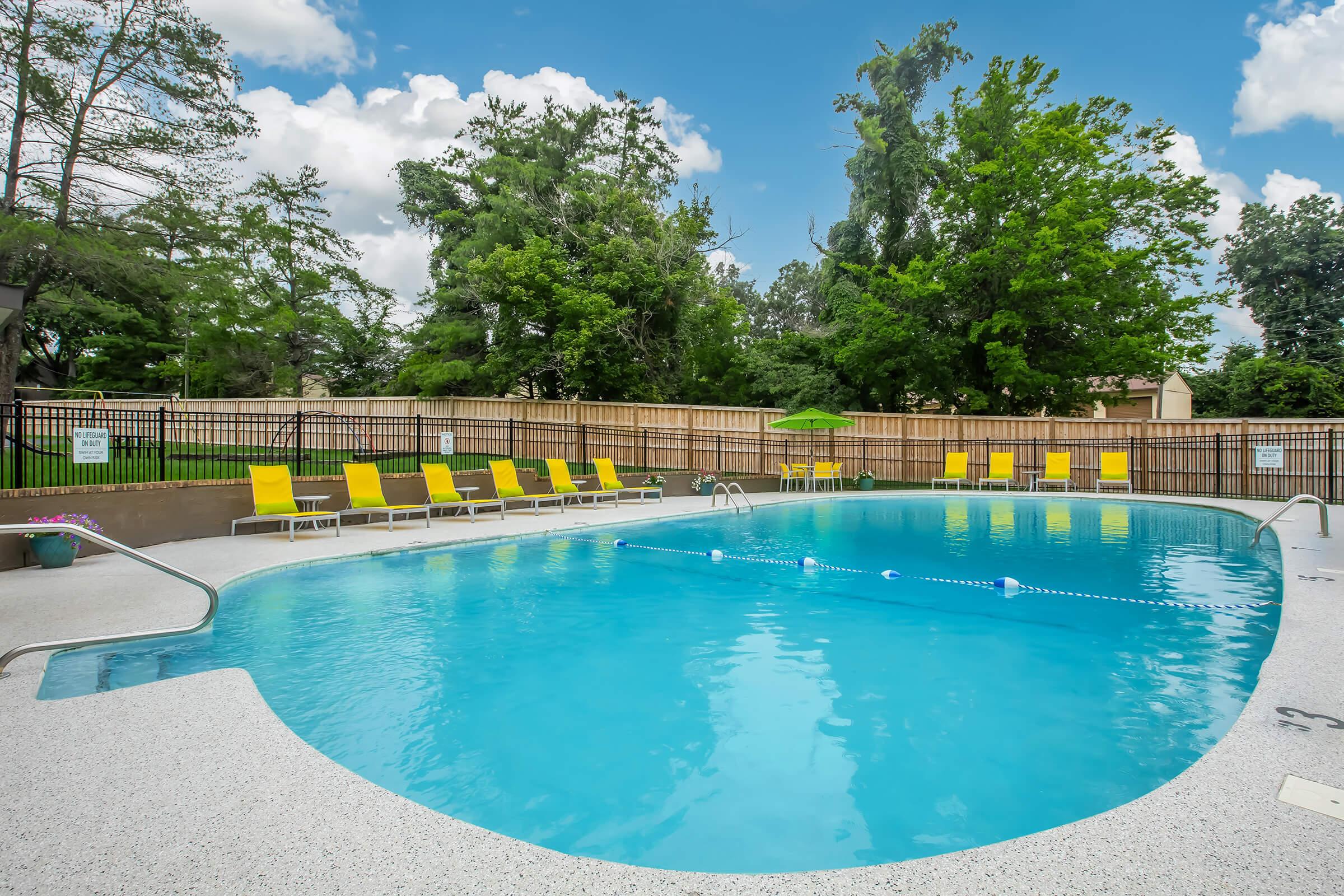 A bright, inviting outdoor swimming pool surrounded by green trees and a wooden fence. Yellow lounge chairs are arranged around the poolside, and there's a shaded area with an umbrella. The sky is partly cloudy, creating a refreshing, relaxing atmosphere.