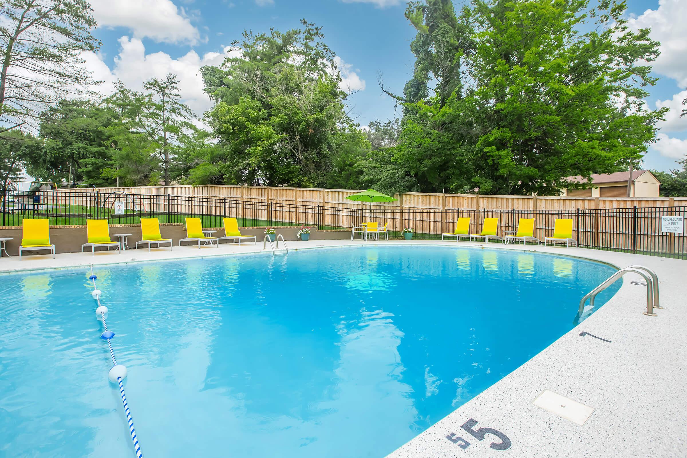 A clear swimming pool surrounded by lounge chairs and greenery, with a wooden fence in the background. The pool area is sunny, featuring a bright blue sky and fluffy clouds. A green umbrella is set up near the edge of the pool, creating a relaxing atmosphere.