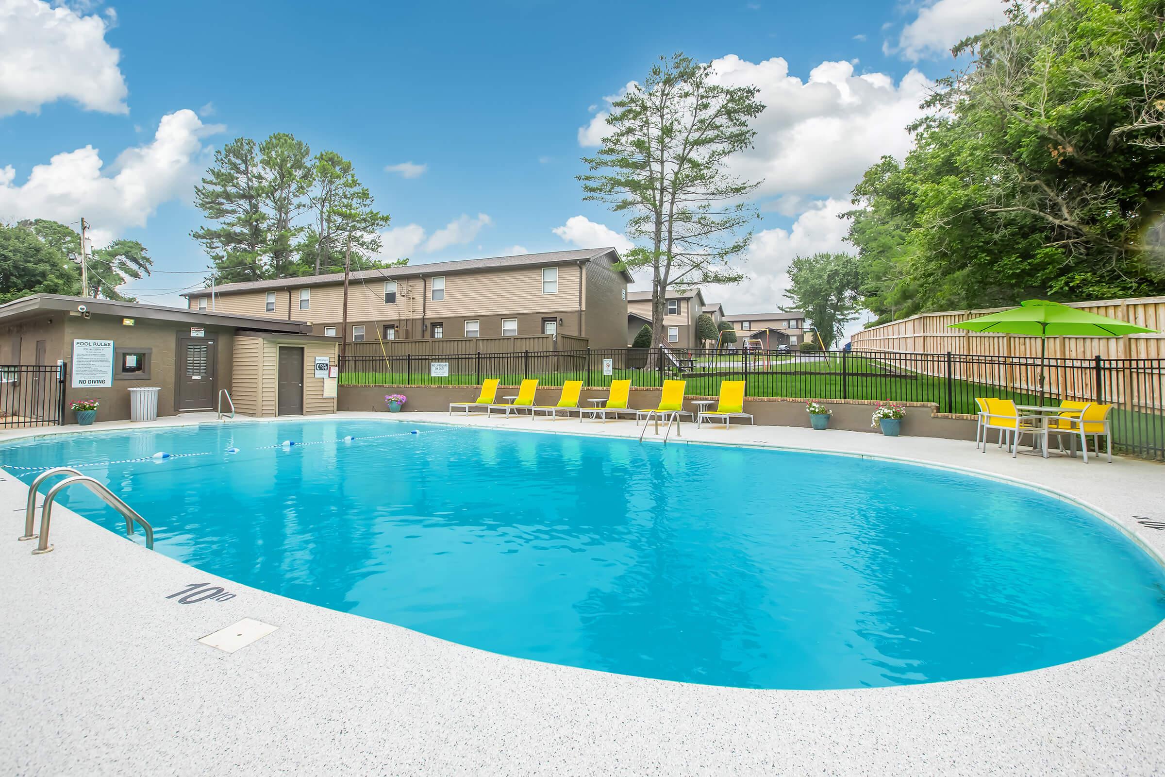A spacious outdoor swimming pool surrounded by lounge chairs and potted plants. In the background, there are apartment buildings and trees under a bright blue sky with fluffy white clouds. The pool area is well-maintained and inviting, perfect for relaxation.