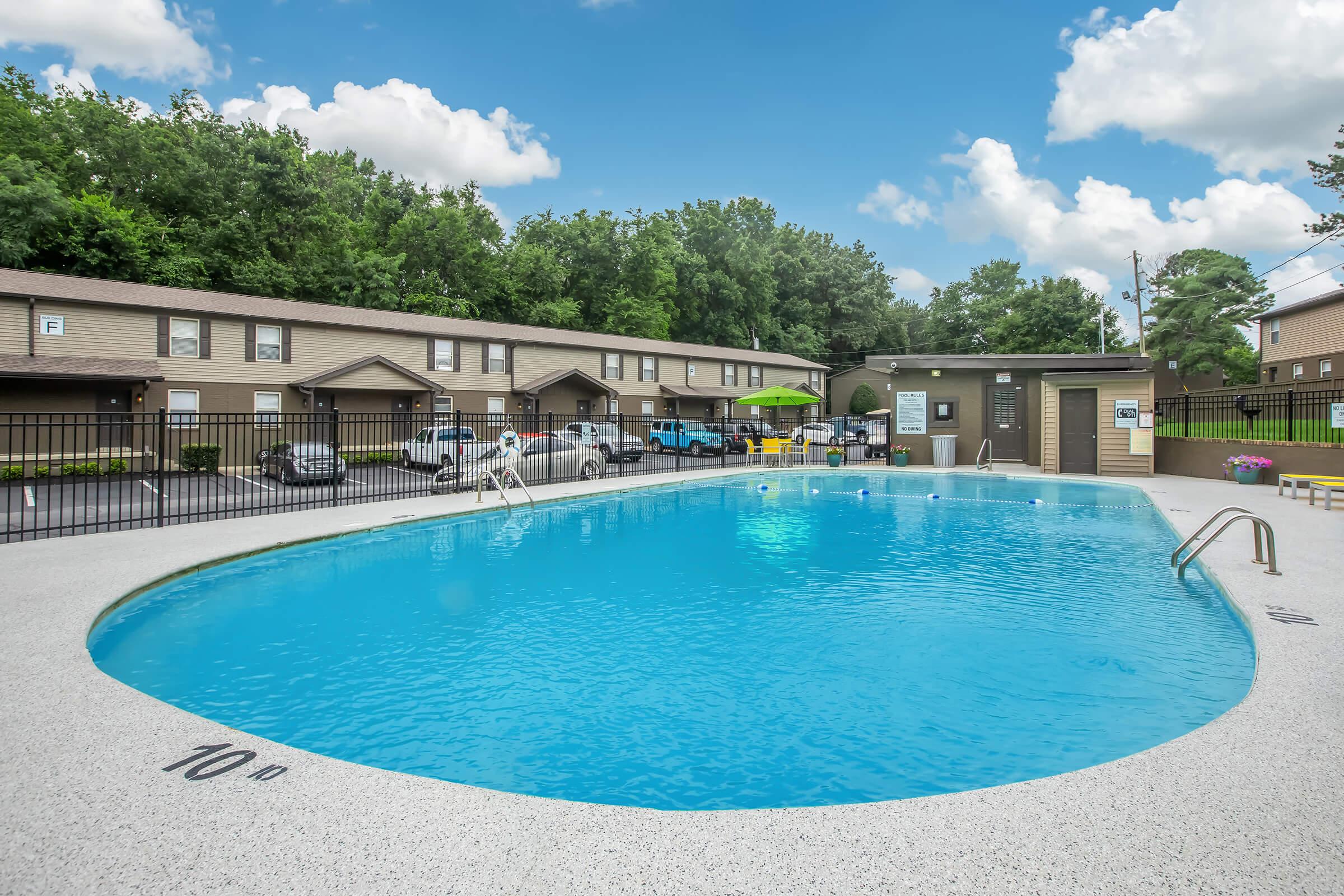 A clear blue swimming pool surrounded by a textured deck, with a ladder leading into the pool. In the background, there are tan apartment buildings with a parking area and a few trees. A lifeguard stands near the pool. Bright clouds and a blue sky complete the scene.