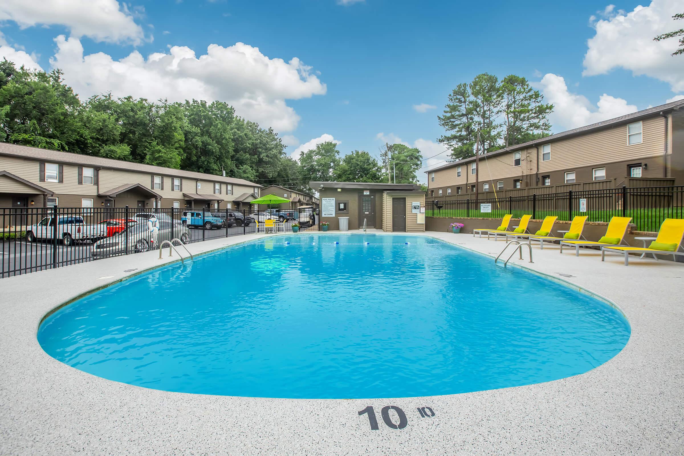 A clear blue swimming pool surrounded by bright yellow lounge chairs, with a few trees in the background. In the distance, there are several tan-colored apartment buildings. The sky is partly cloudy, creating a vibrant and inviting atmosphere.