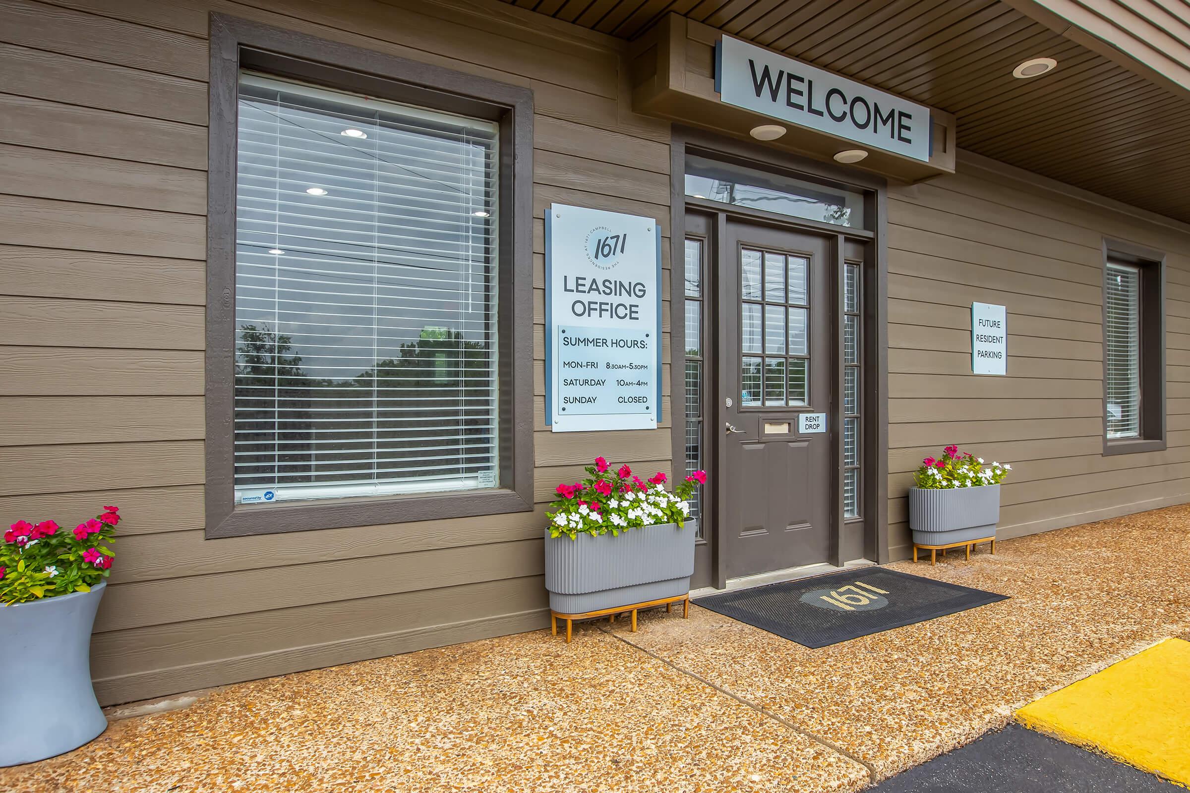 Entrance to a leasing office with a "Welcome" sign above the door. The building features brown wooden siding, large windows, and potted flowers flanking the entrance. A sign lists the office name, hours of operation, and contact information. The area is well-maintained and inviting.