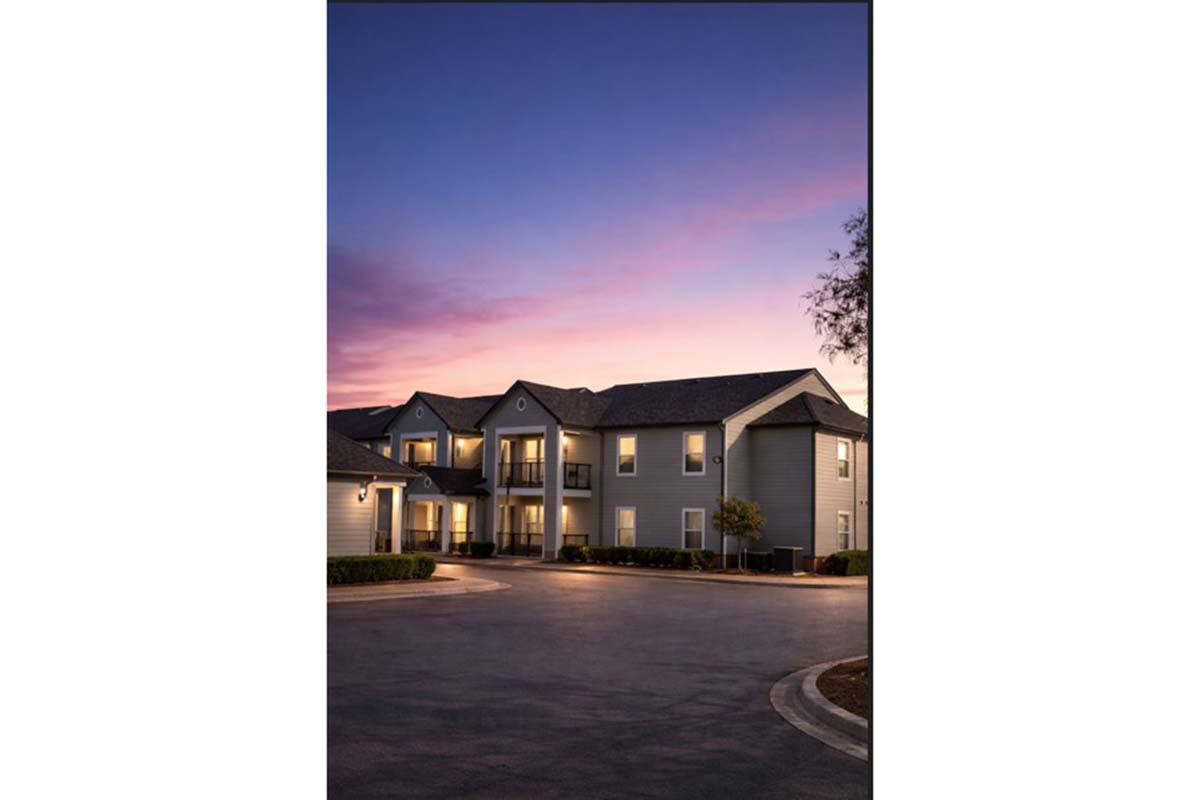 A modern apartment building at dusk, featuring multiple balconies and warm lights illuminating the exterior. The sky above showcases a gradient of colors from orange to purple, creating a serene atmosphere. Trees and shrubs are visible in the surroundings, enhancing the residential setting.