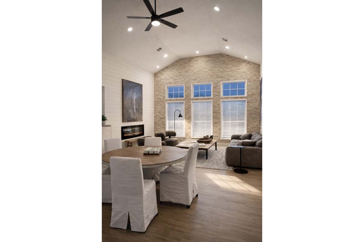 A spacious modern living room featuring a vaulted ceiling, large windows with white shutters, and a cozy seating area with a sectional sofa and a coffee table. A round dining table with white chairs is visible, along with a modern ceiling fan and an electric fireplace in the background.