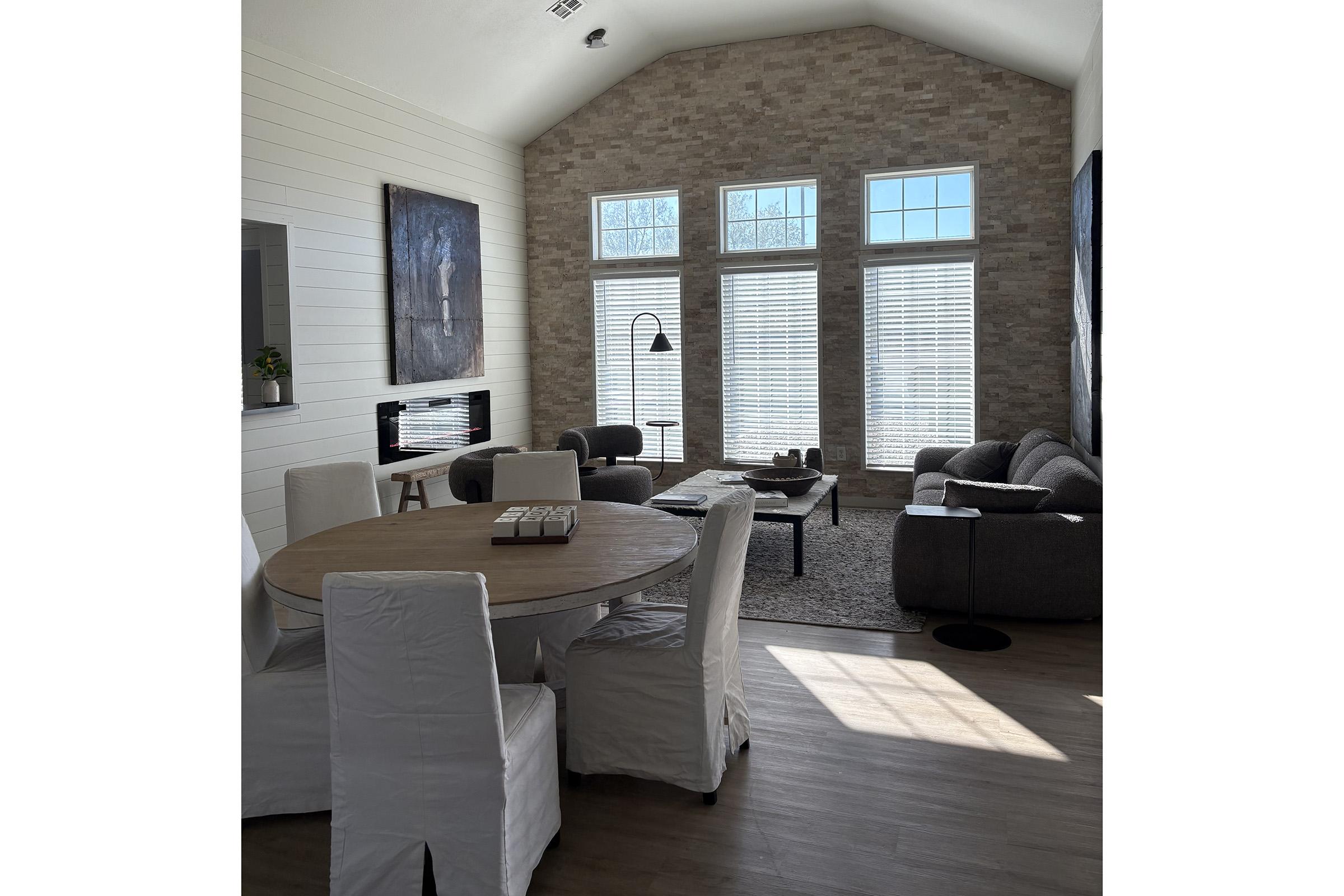 A bright and spacious living area featuring large windows that let in natural light. There's a round wooden dining table with white chairs, a cozy gray sofa, and a coffee table in the sitting area. The walls are adorned with artwork, and a sleek floor lamp adds a modern touch.