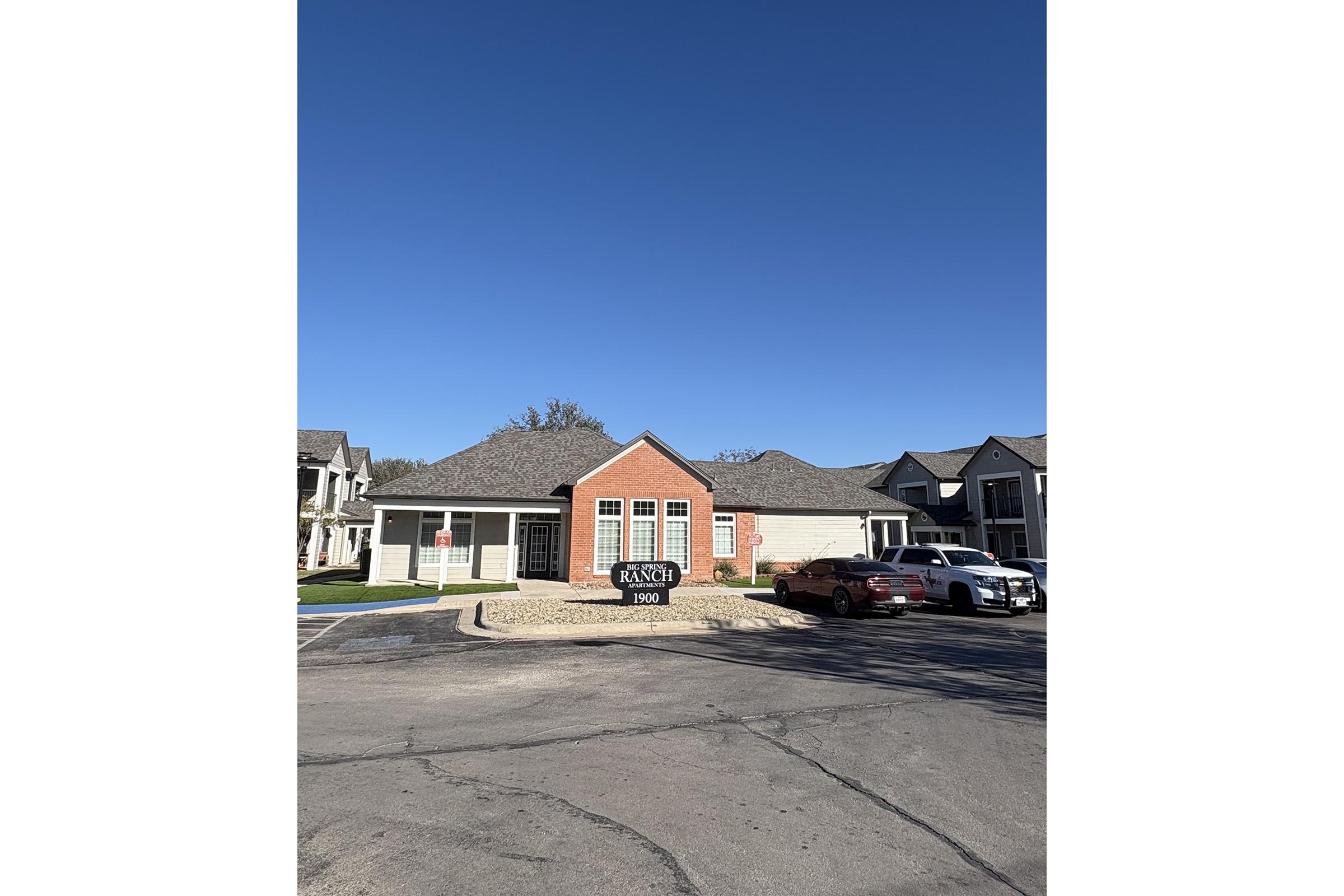 A modern building with a red brick entryway and large windows, surrounded by a landscaped area. In the foreground, there is a paved parking lot with two parked cars. A clear blue sky is visible above, and additional buildings can be seen to the right.