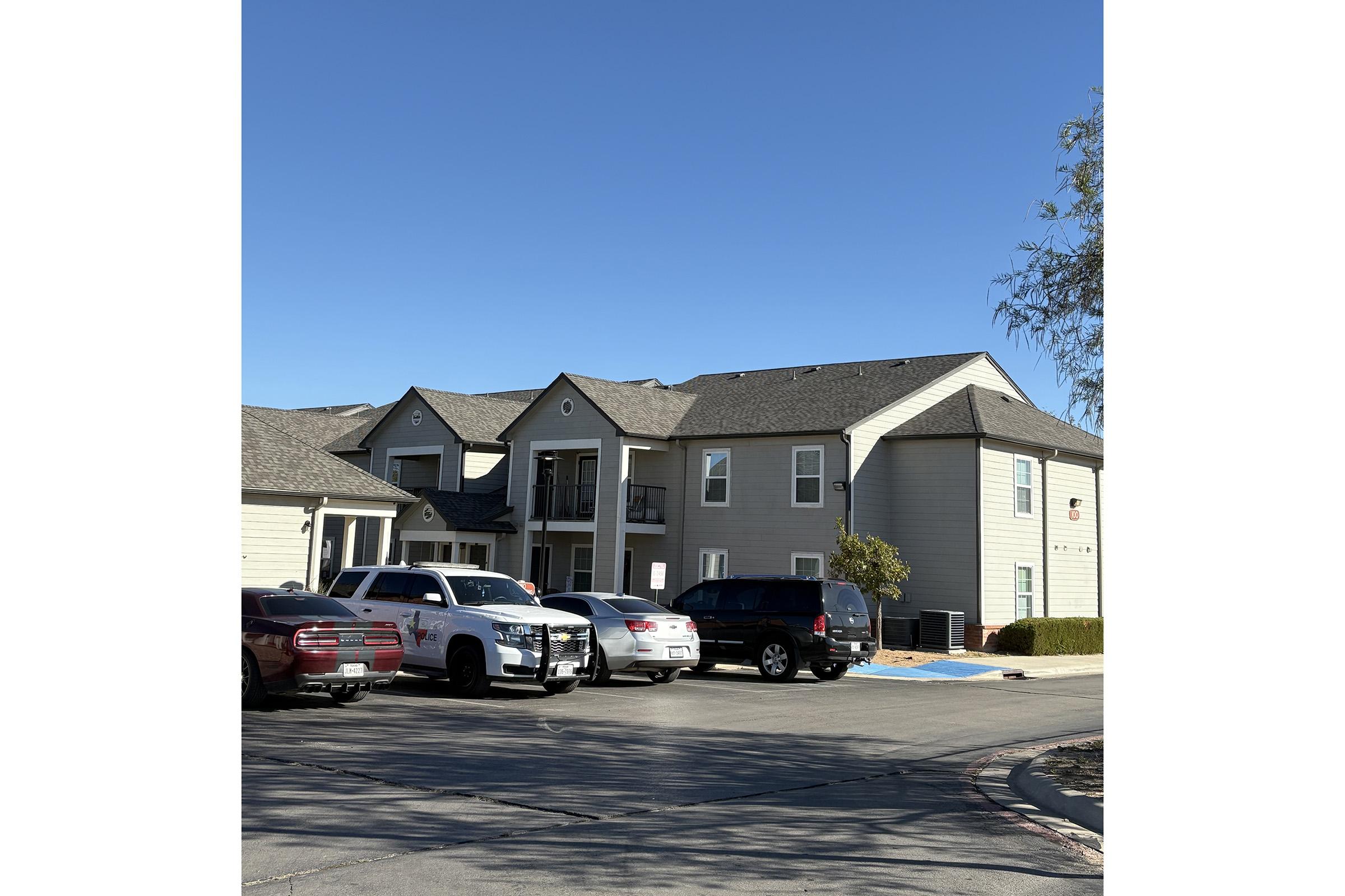 A two-story apartment building with a mix of gray and beige siding. In front, there are several parked cars, including a police vehicle. The sky is clear and blue, and there are some trees nearby. The building has balconies, and the area appears to be well-maintained.