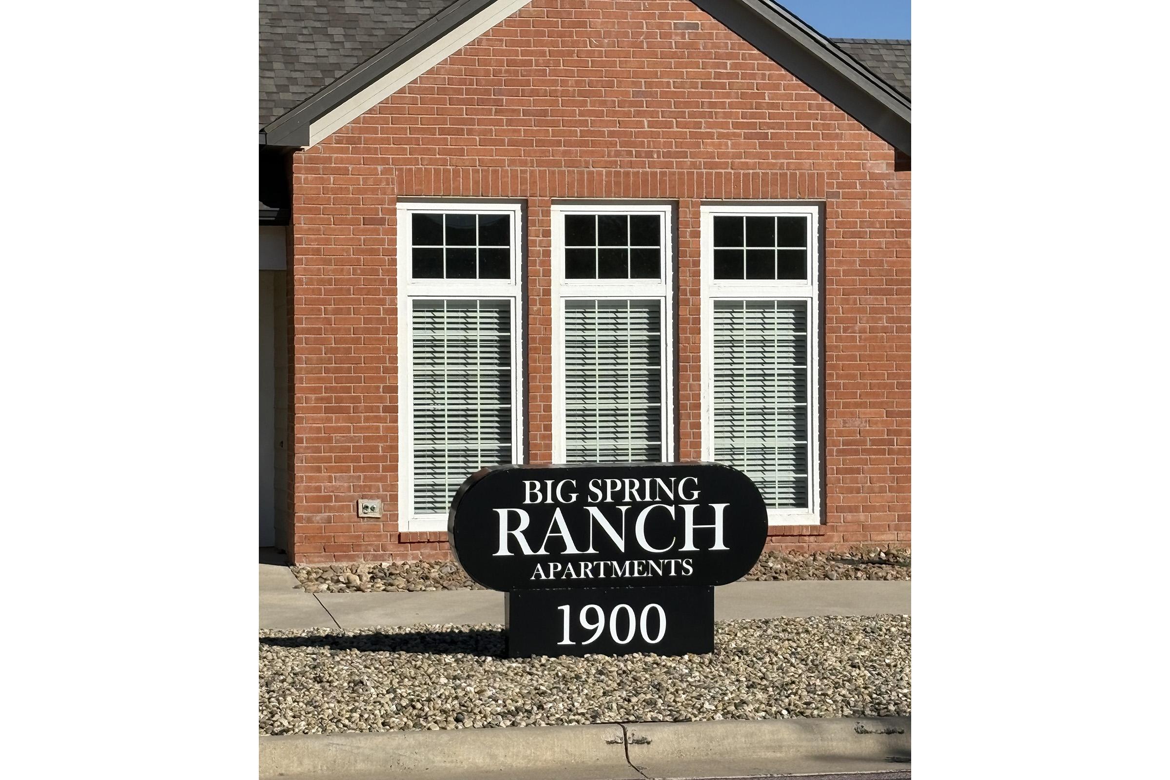 Sign for Big Spring Ranch Apartments located at 1900, in front of a brick building with multiple windows and shutters. The sign is black with white text, emphasizing the name of the apartments. The surrounding area features small rocks and landscaping.