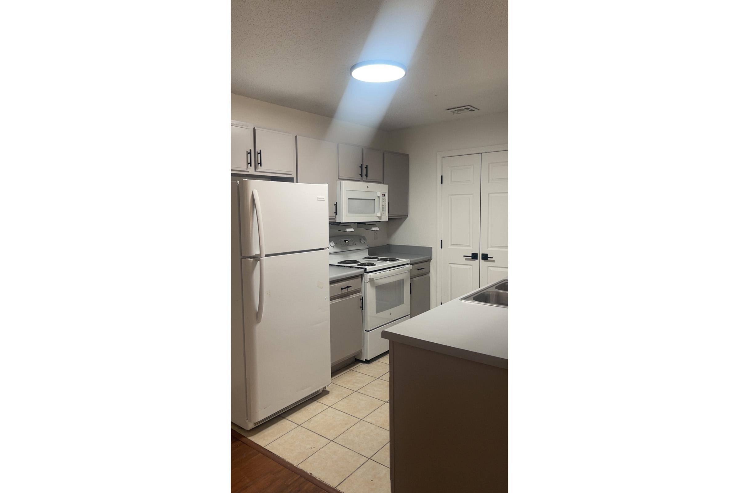 Modern kitchen with light-colored cabinets, a white refrigerator, a white microwave above an electric stove, and a silver sink on an island. The tiled floor complements the appliances, and there is a ceiling light providing illumination. The overall look is clean and functional.