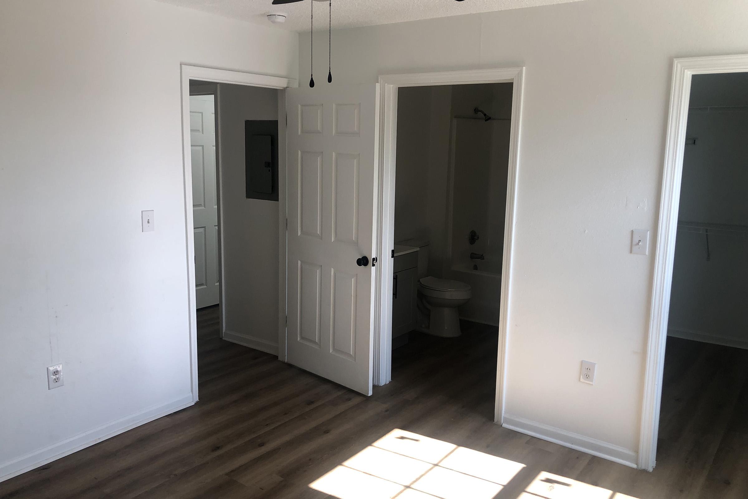 A well-lit room featuring a light-colored wall and wood-like flooring. There are three doors: one leading to a bathroom with a toilet visible, and the other two opening to adjacent spaces. Natural light is streaming in from a window, creating a warm atmosphere.