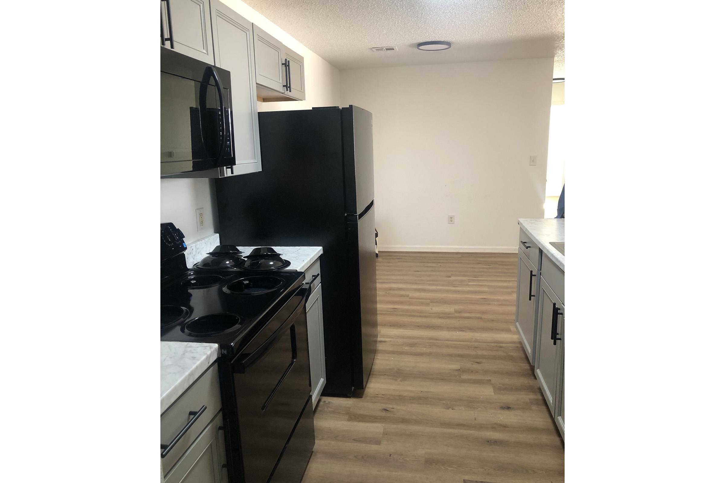 Modern kitchen with light gray cabinets, black appliances including a stove and refrigerator, and a white countertop. The flooring is a light wood laminate. A spacious area is seen at the end of the kitchen, leading to a well-lit room with a window.