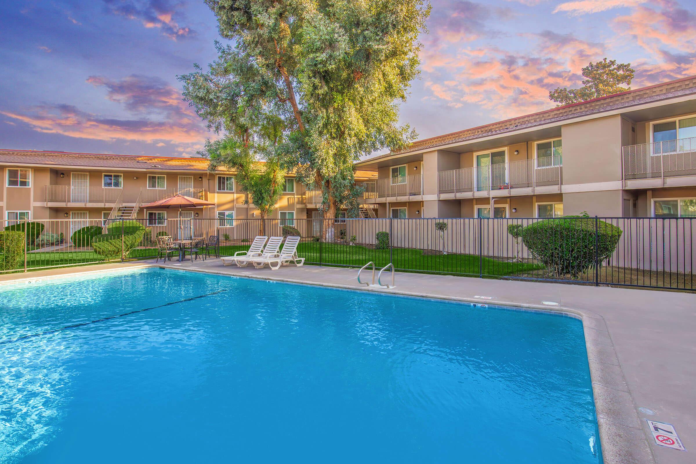 A clear blue swimming pool surrounded by lounge chairs, with well-manicured grass and bushes. In the background, there are two-story apartments with balconies and a vibrant sunset sky, creating a relaxing and inviting atmosphere.