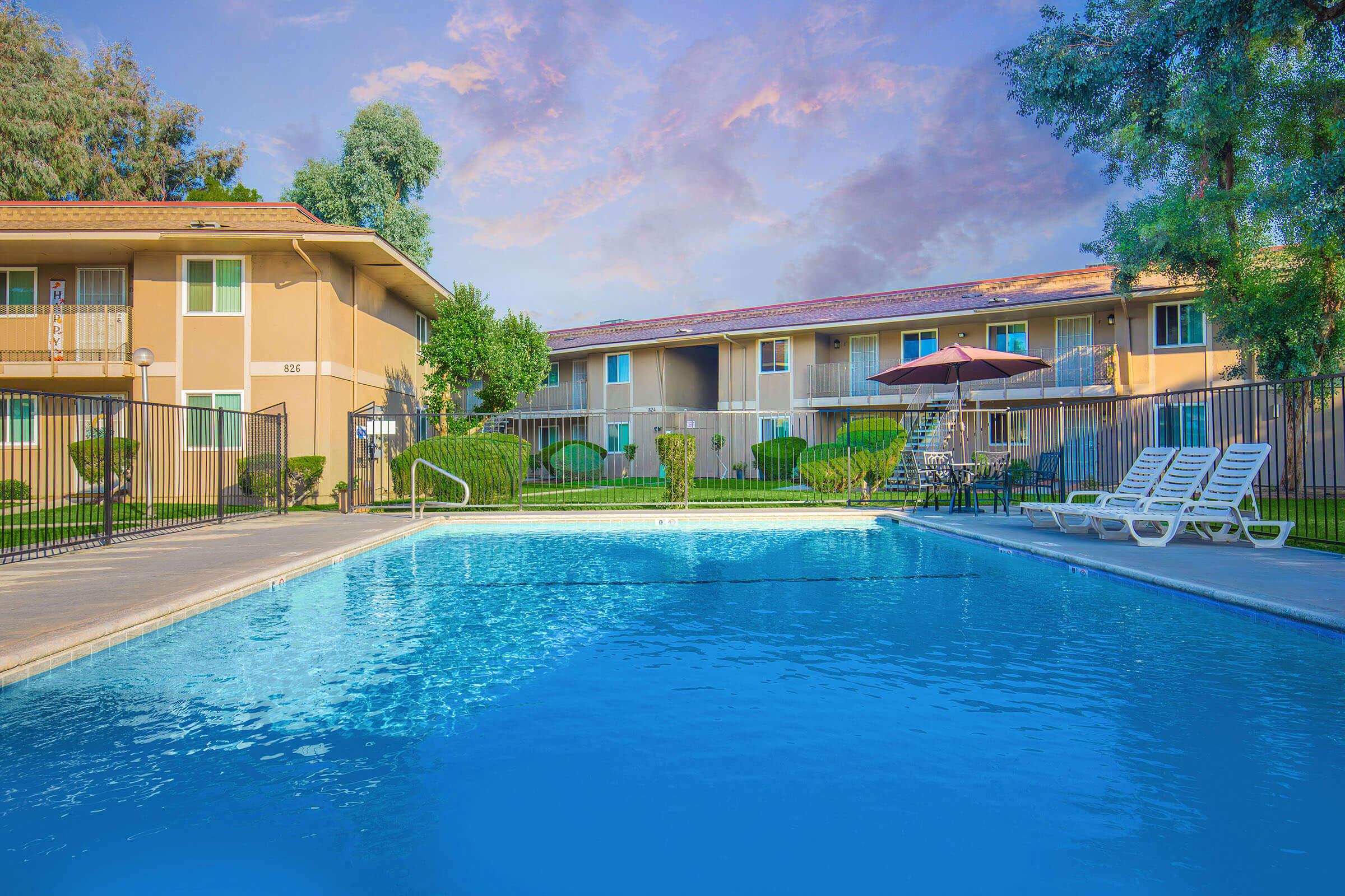 A clean, inviting swimming pool surrounded by lounge chairs, near two-story apartment buildings with landscaped greenery. An umbrella provides shade on the pool deck, and the sky is partly cloudy with a warm sunset glow. Perfect setting for relaxation and outdoor enjoyment.