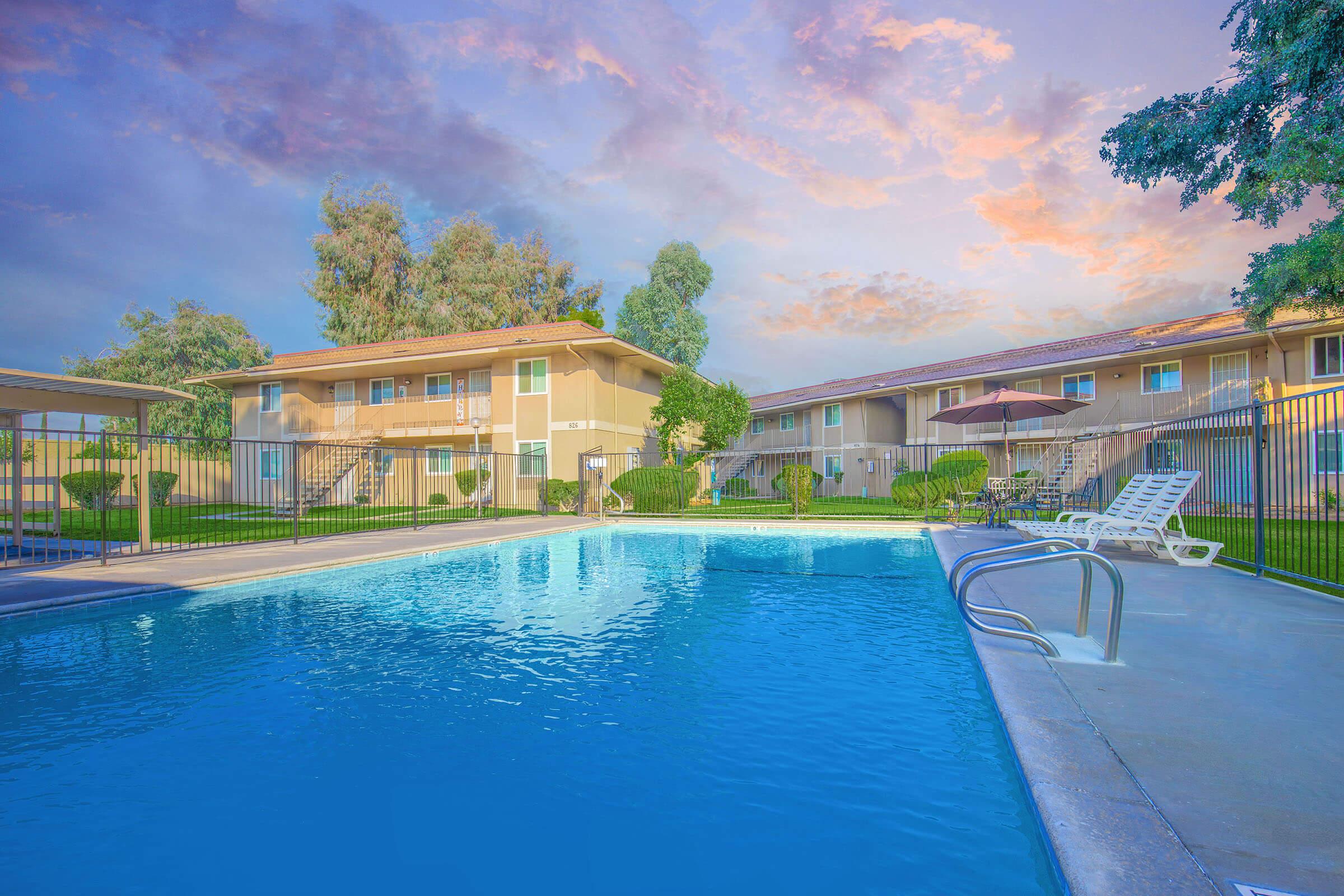 A tranquil swimming pool in front of an apartment complex, surrounded by green lawns and trees. The sky features soft pastel hues during sunset, and a few lounge chairs are placed by the poolside. There is a covered seating area and an umbrella visible, creating a relaxing outdoor atmosphere.