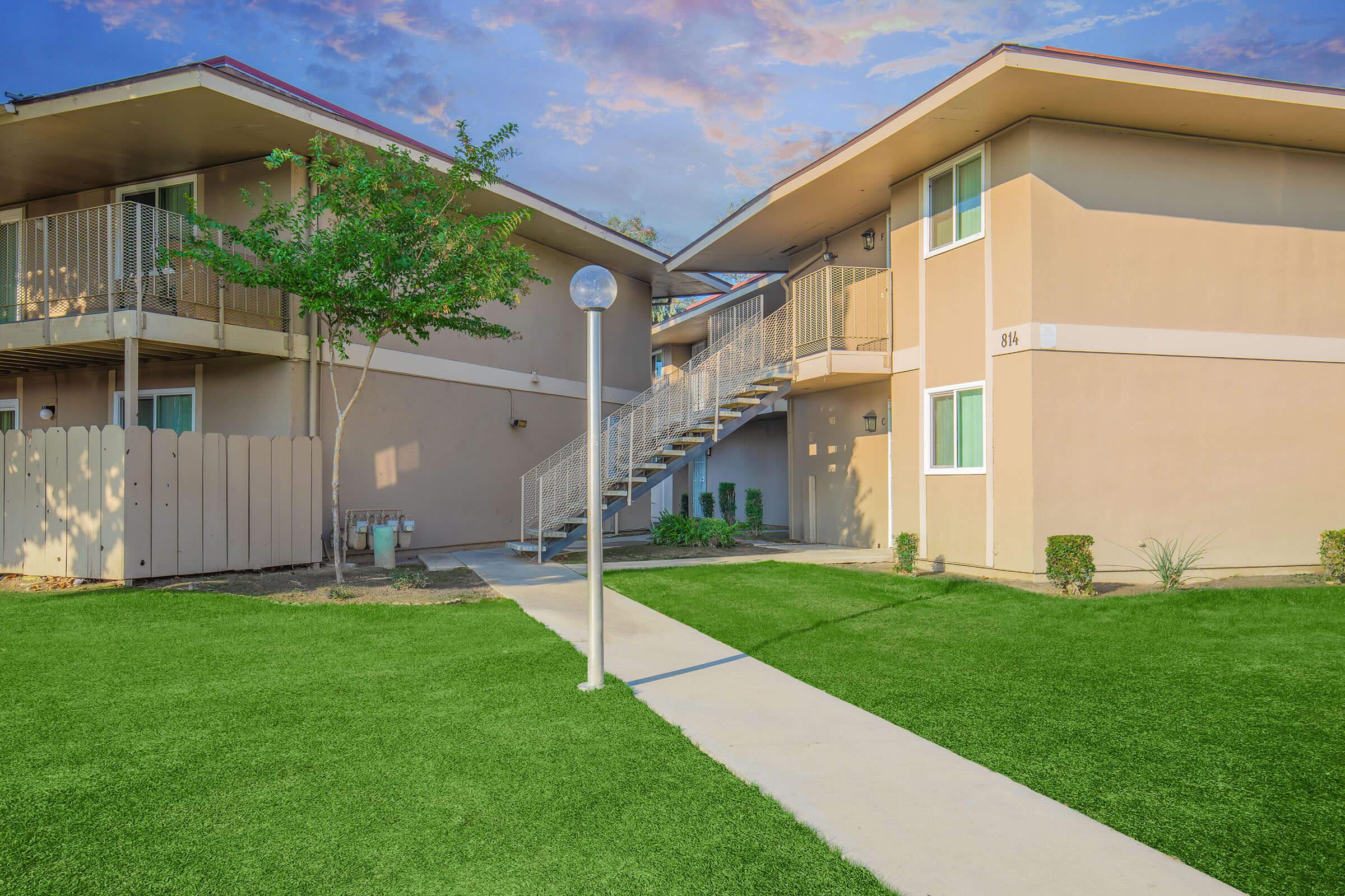 A well-maintained apartment complex featuring two buildings with beige exteriors, surrounded by lush green grass. A pathway leads through the complex, with a lamppost and small shrubs enhancing the landscaping. A staircase connects the upper levels of the buildings, creating a welcoming atmosphere.