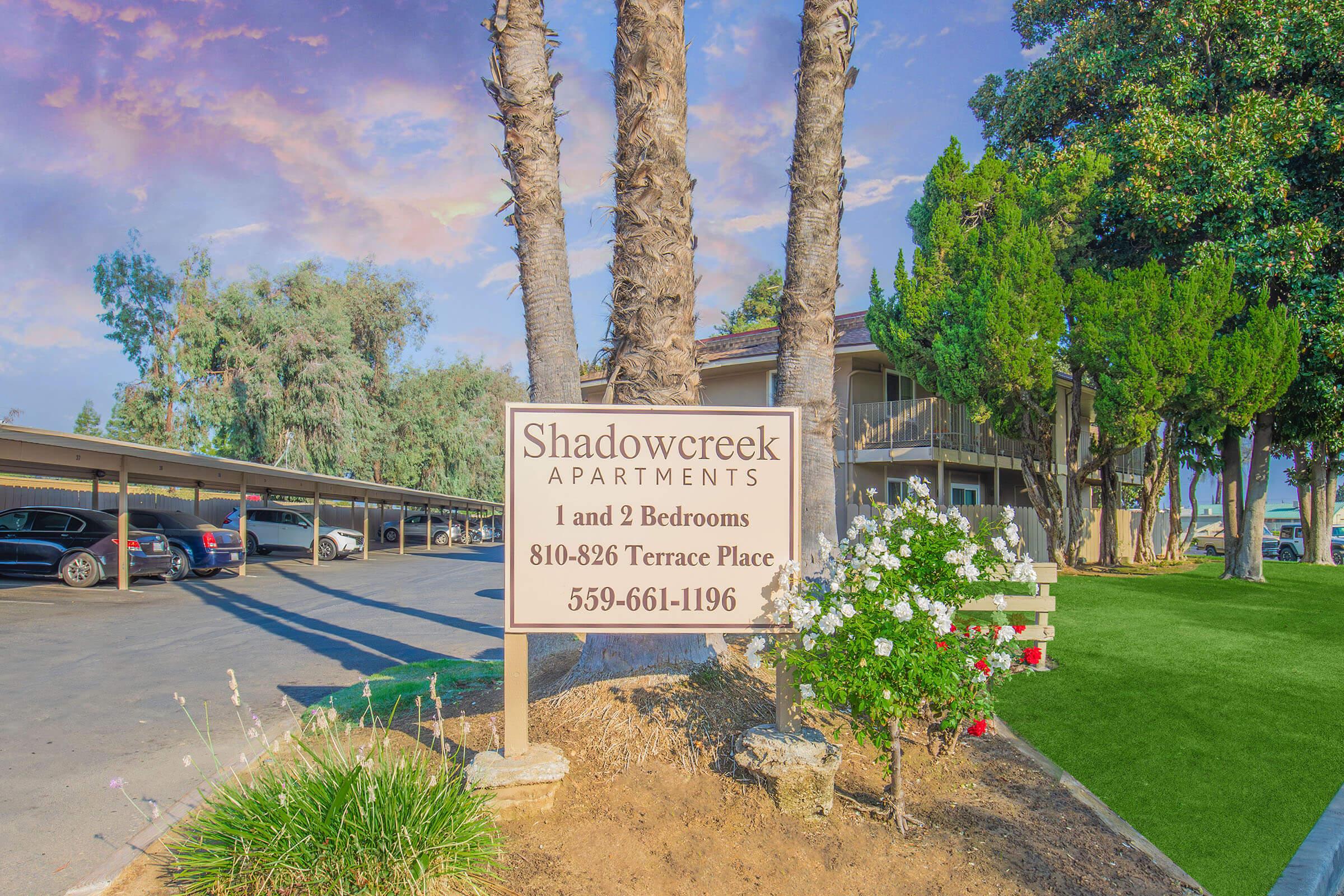 Sign for Shadowcreek Apartments featuring contact information and details about available 1 and 2-bedroom units. The sign is surrounded by trees and flowers, with a parking area visible in the background. Clear blue sky adds to the inviting atmosphere of the apartment complex.