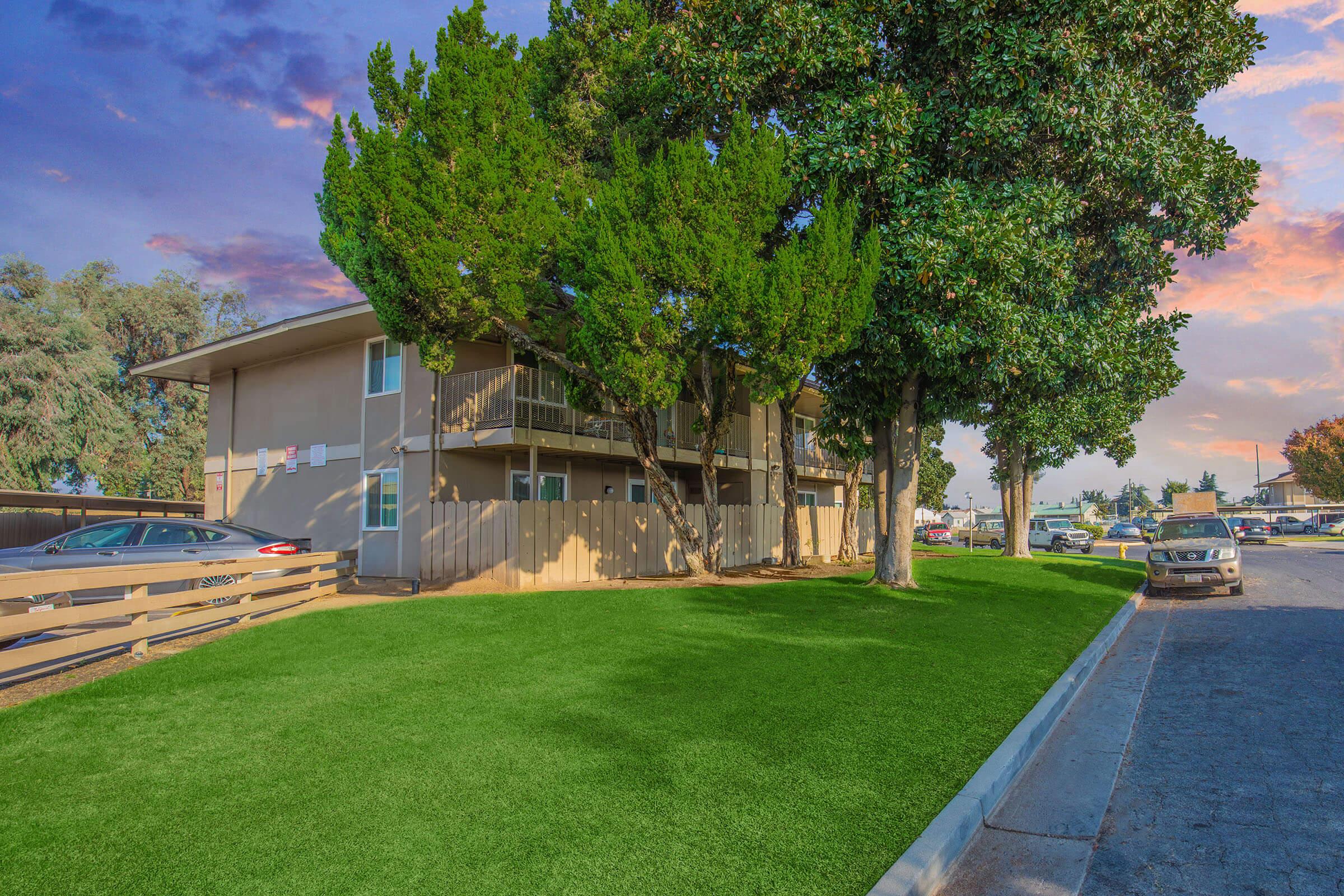 A modern apartment building surrounded by lush green grass and trees, with vehicles parked nearby. The scene is set against a colorful sunset sky, creating a serene and inviting atmosphere.
