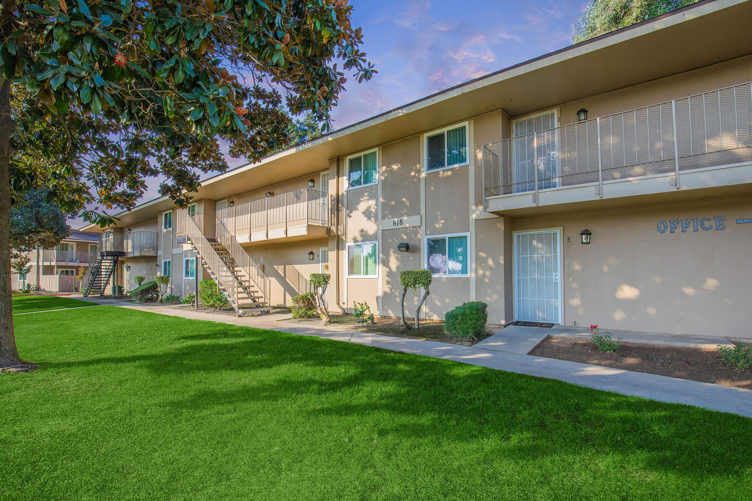 Two-story apartment building with multiple units, featuring a staircase leading to the second floor. The area is well-maintained with green grass, shrubs, and trees. The facade includes an office entrance with large windows and a door, set against a clear blue sky.