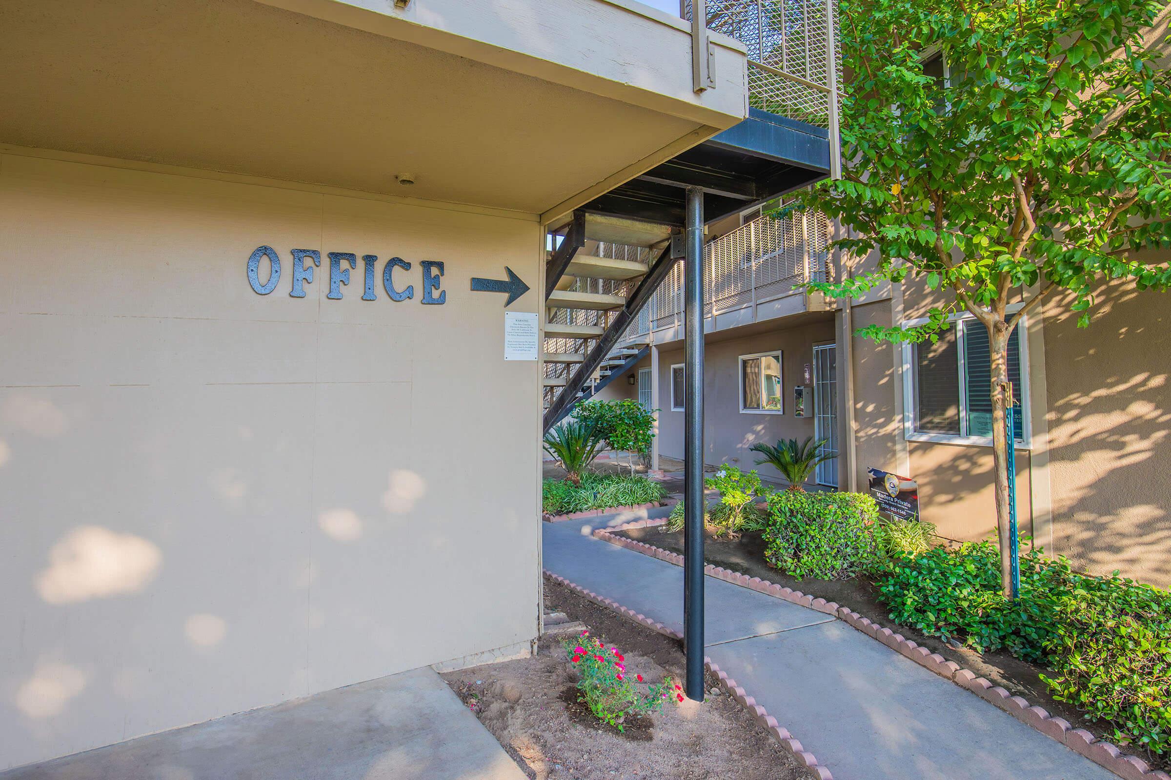 A pathway leading to an office area, marked by a sign pointing left that reads "OFFICE." The scene features well-maintained landscaping with small plants and flowers, and a staircase to the right. The building is part of an apartment complex, with sunlight filtering through the trees.