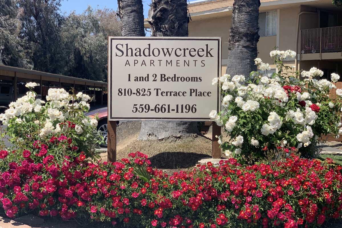Sign for Shadowcreek Apartments featuring information about available 1 and 2-bedroom units. The sign is framed by colorful flowers, including white and red roses, in front of palm trees and an apartment building. Contact number displayed is 559-661-1196 and the address is 810-825 Terrace Place.