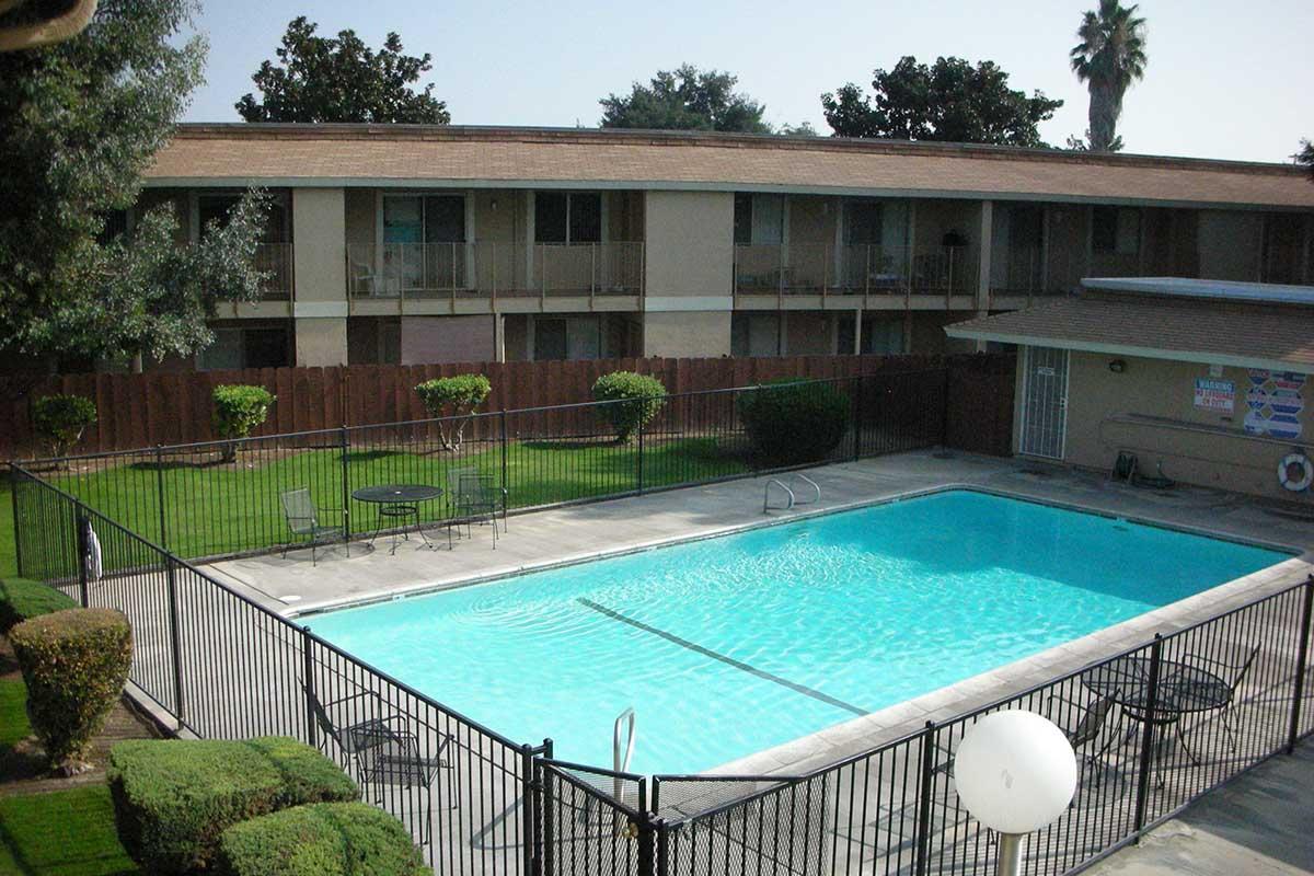 A clear blue swimming pool surrounded by a black metal fence, with nearby lounge chairs and small tables. In the background, a two-story building with multiple balconies and well-maintained greenery. The scene is bright and sunny, indicating a warm day.