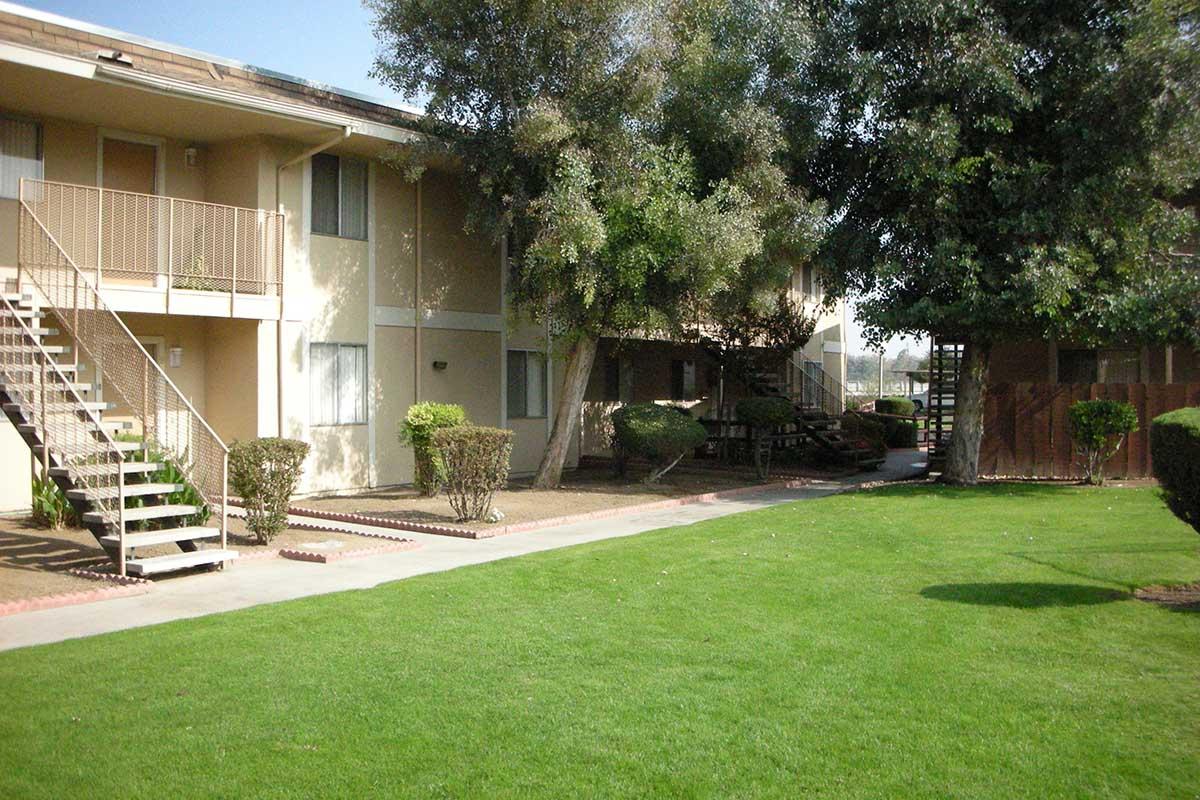 A residential area featuring two-story apartment buildings with outdoor staircases. The landscape includes well-maintained grassy areas, shrubs, and a pathway leading through the greenery. The atmosphere is peaceful and inviting, with clear blue skies above.