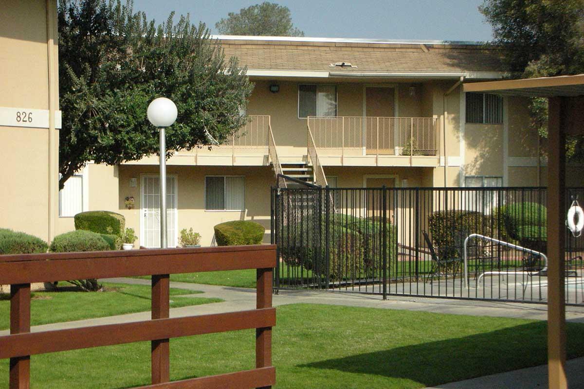A residential apartment complex featuring two levels with balconies, surrounded by manicured lawns and shrubs. A gated area with a swimming pool is visible in the foreground, along with a wooden railing leading to the entrance. The setting is sunny with clear blue skies.