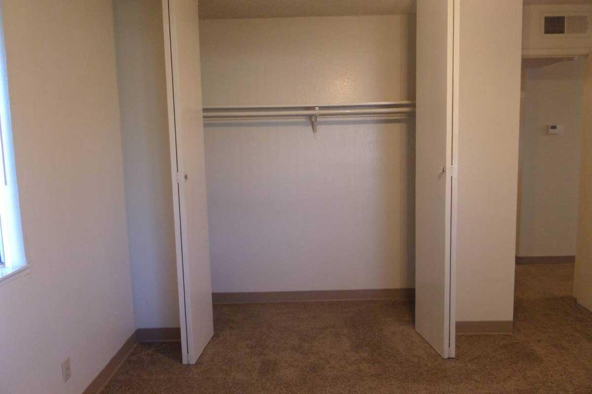 An empty room featuring two open closet doors revealing a simple hanging rod. The walls are beige, and the carpet is a light brown color. There is a window with natural light on the left and a doorway visible on the right, suggesting an open layout.
