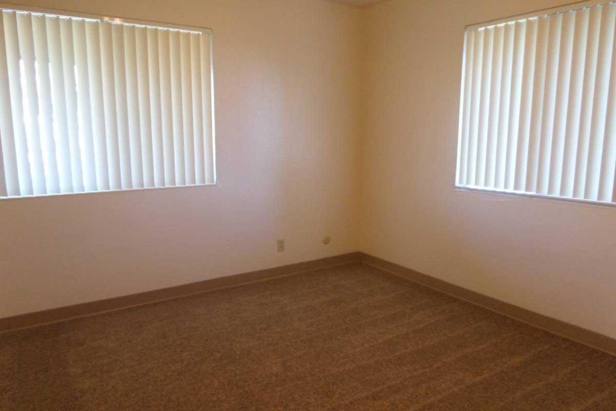 Empty room with beige walls and brown carpet. There are two windows with vertical blinds letting in natural light. The overall space appears clean and uncluttered, suitable for various uses such as an office or bedroom.