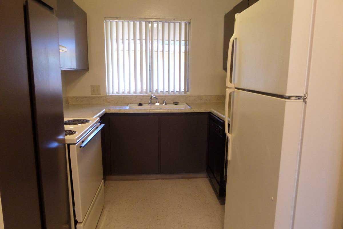 Compact kitchen interior showcasing a refrigerator, stove, and sink. Below the window, there are countertops with dark cabinetry, complemented by light-colored walls. The space features vertical blinds letting in natural light, creating a functional and inviting cooking area.