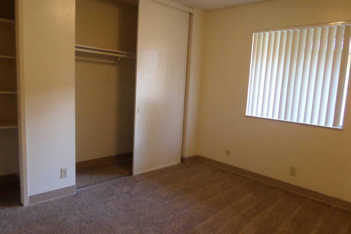 A vacant room with beige walls, a window with vertical blinds, and light brown carpet. There is a closet with sliding doors and a shelf inside, providing storage space. The overall ambiance is simple and uncluttered, suitable for personal customization.