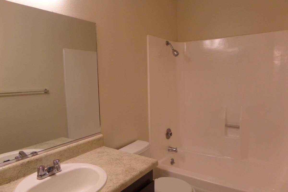 A clean bathroom featuring a white bathtub with a showerhead, a round sink with a faucet, and a large mirror above the sink. The walls are a light color, and there is a towel rack on the wall. The overall appearance is tidy and minimalistic.
