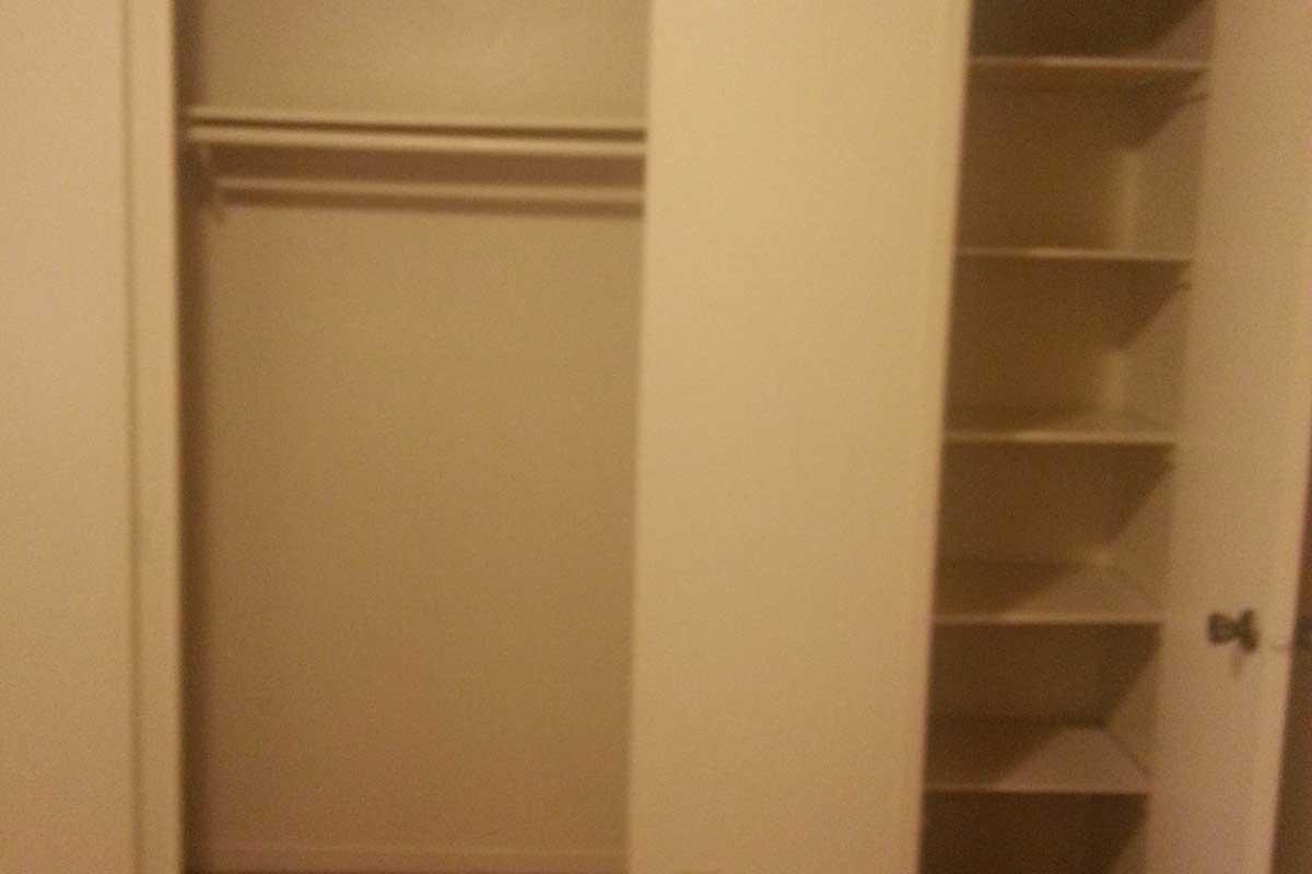 A partially open closet with one side showing a hanging rod and the other side featuring several empty shelves. The walls and shelves are painted a neutral color. The floor is visible at the bottom, and the closet door is slightly ajar.