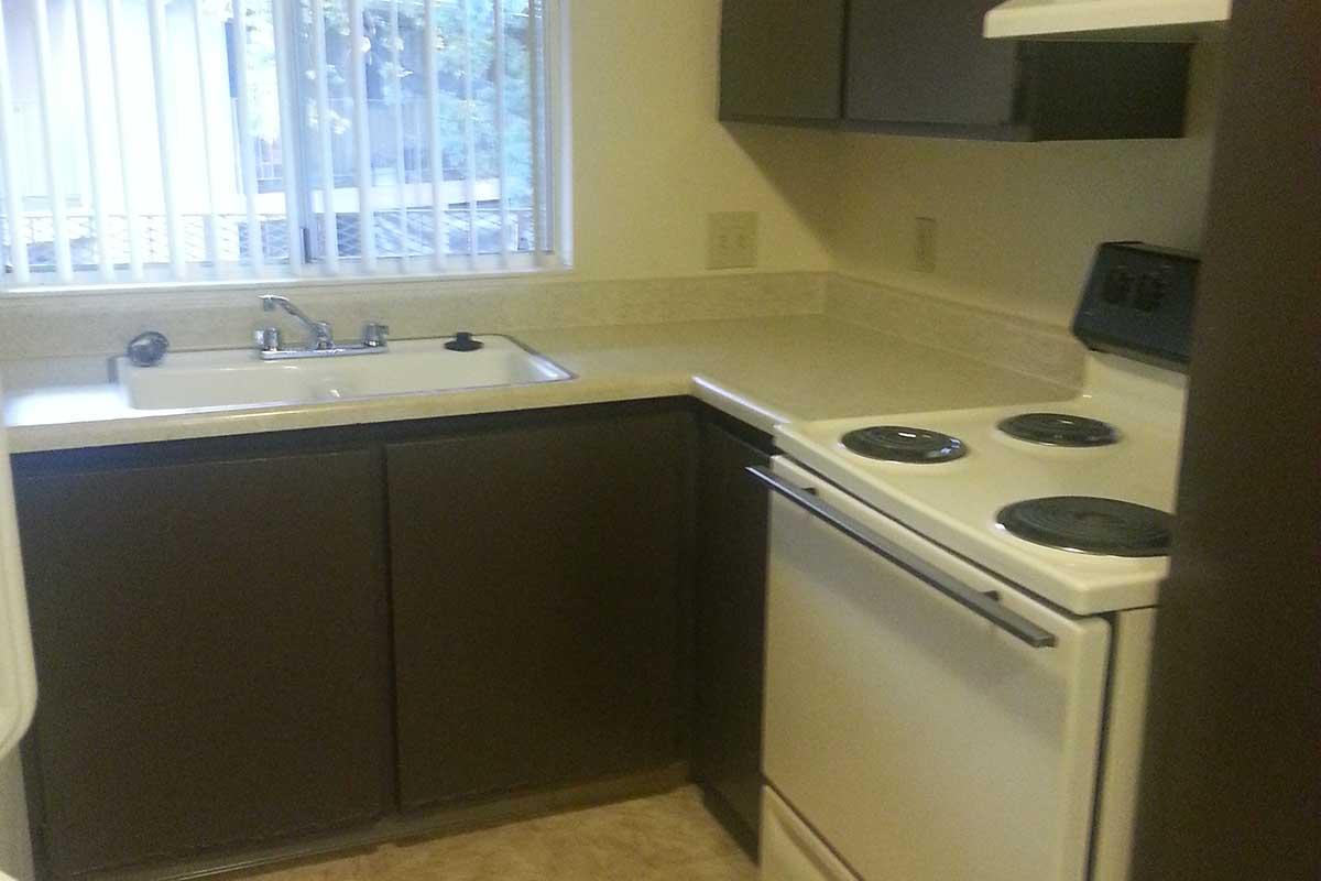 A small kitchen featuring a white stove and oven, a sink with a faucet, and dark cabinets. The countertop is light-colored, and there are window blinds partially drawn, allowing natural light to enter. The flooring is a light tile, complementing the overall neutral color scheme.