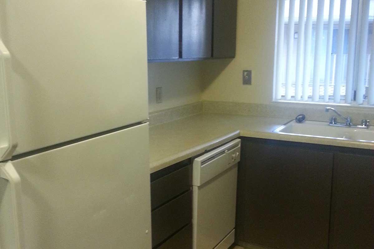 A modern kitchen featuring white appliances, including a refrigerator and dishwasher. The countertop is light-colored with a sleek finish, and there are dark wooden cabinets above and below. A window with vertical blinds provides natural light to the space.