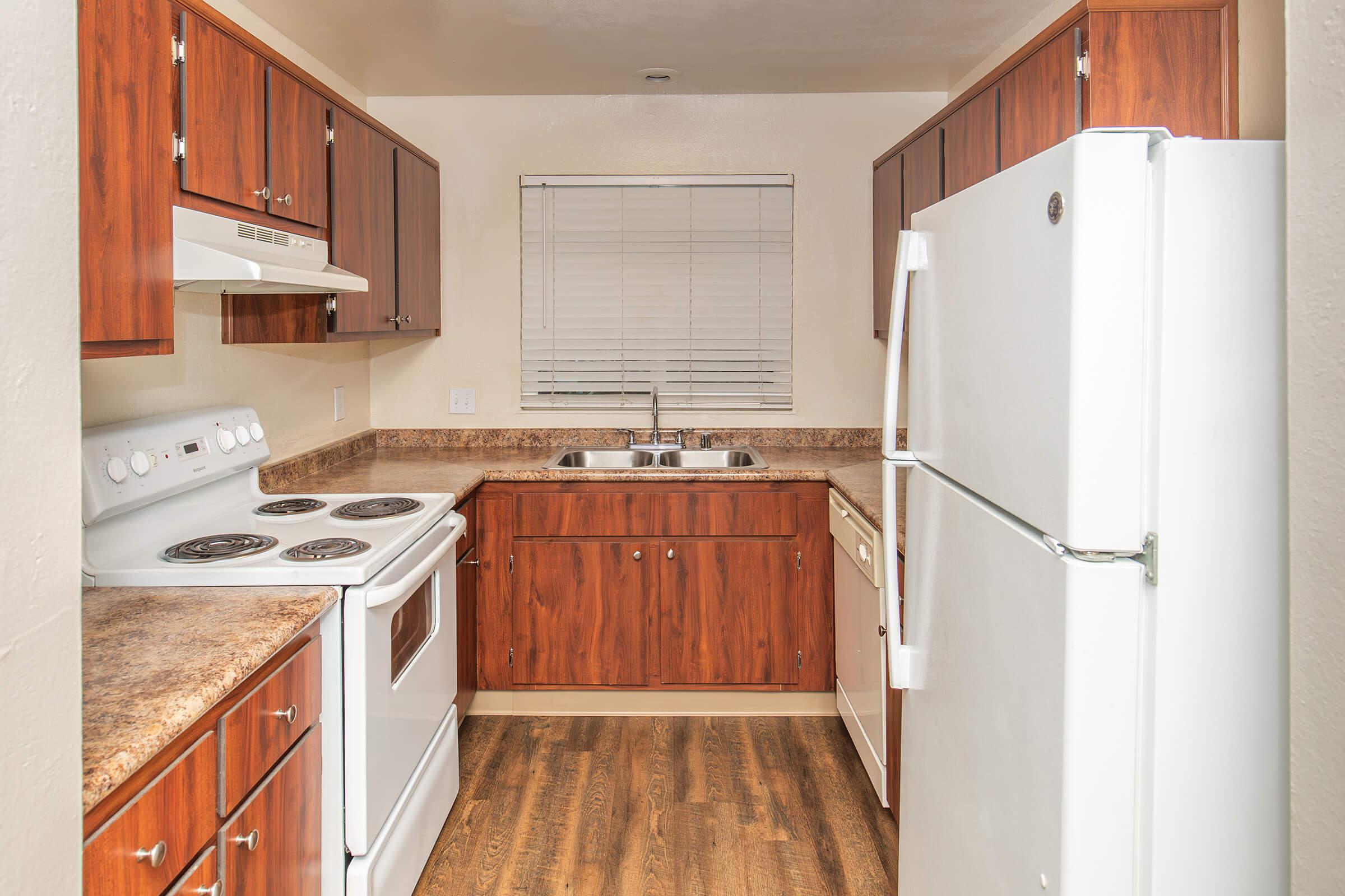 A well-lit kitchen featuring wooden cabinetry, a white refrigerator, an oven with a stovetop, and a double sink. The countertops are brown and there is a window with blinds allowing natural light into the space. The floor is finished with a wood-like texture.