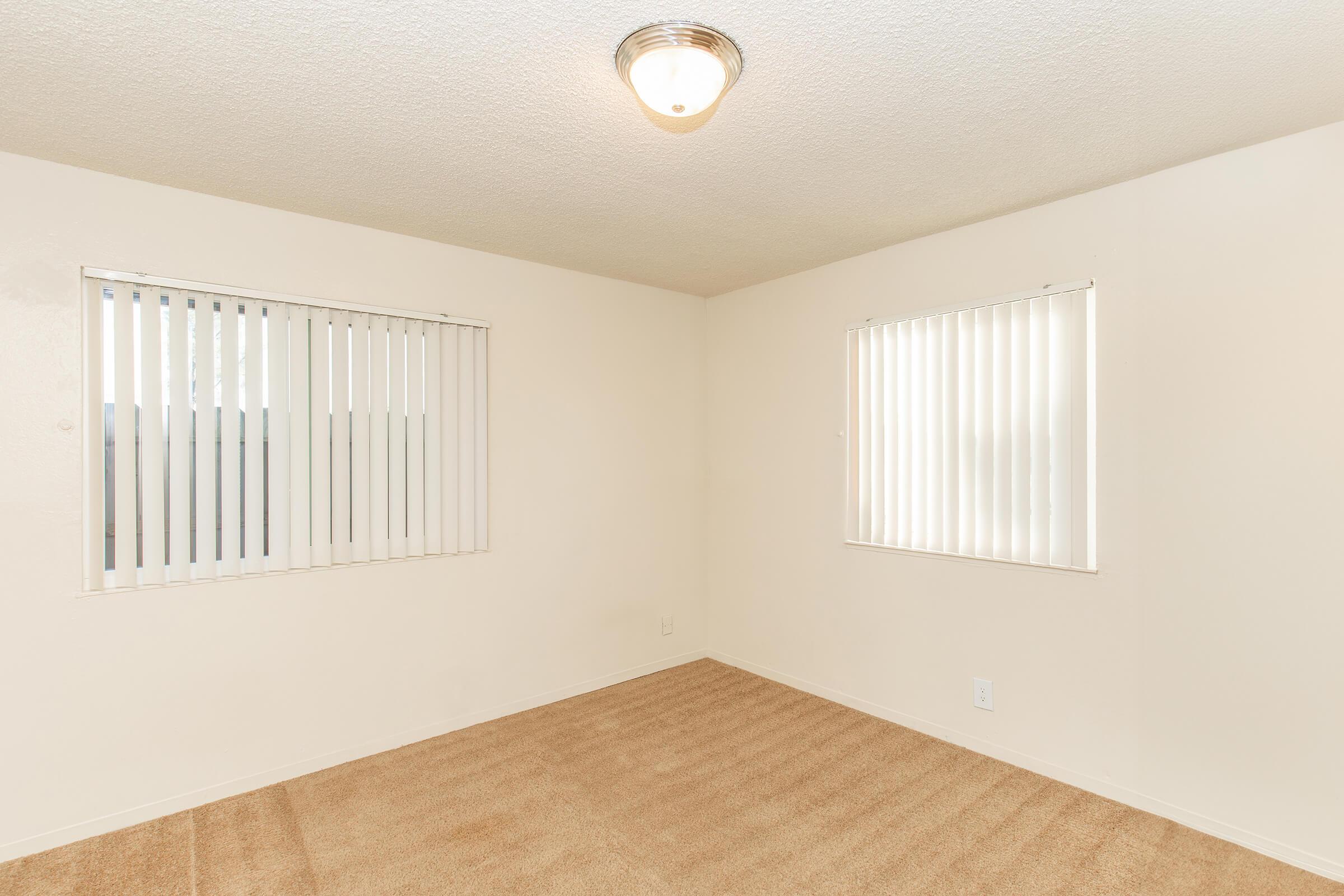 A vacant room with light beige walls and carpet. It features two windows with vertical blinds allowing natural light to enter. A ceiling light fixture is centered in the room, creating a bright and airy atmosphere. The space appears clean and well-maintained, ideal for various uses.