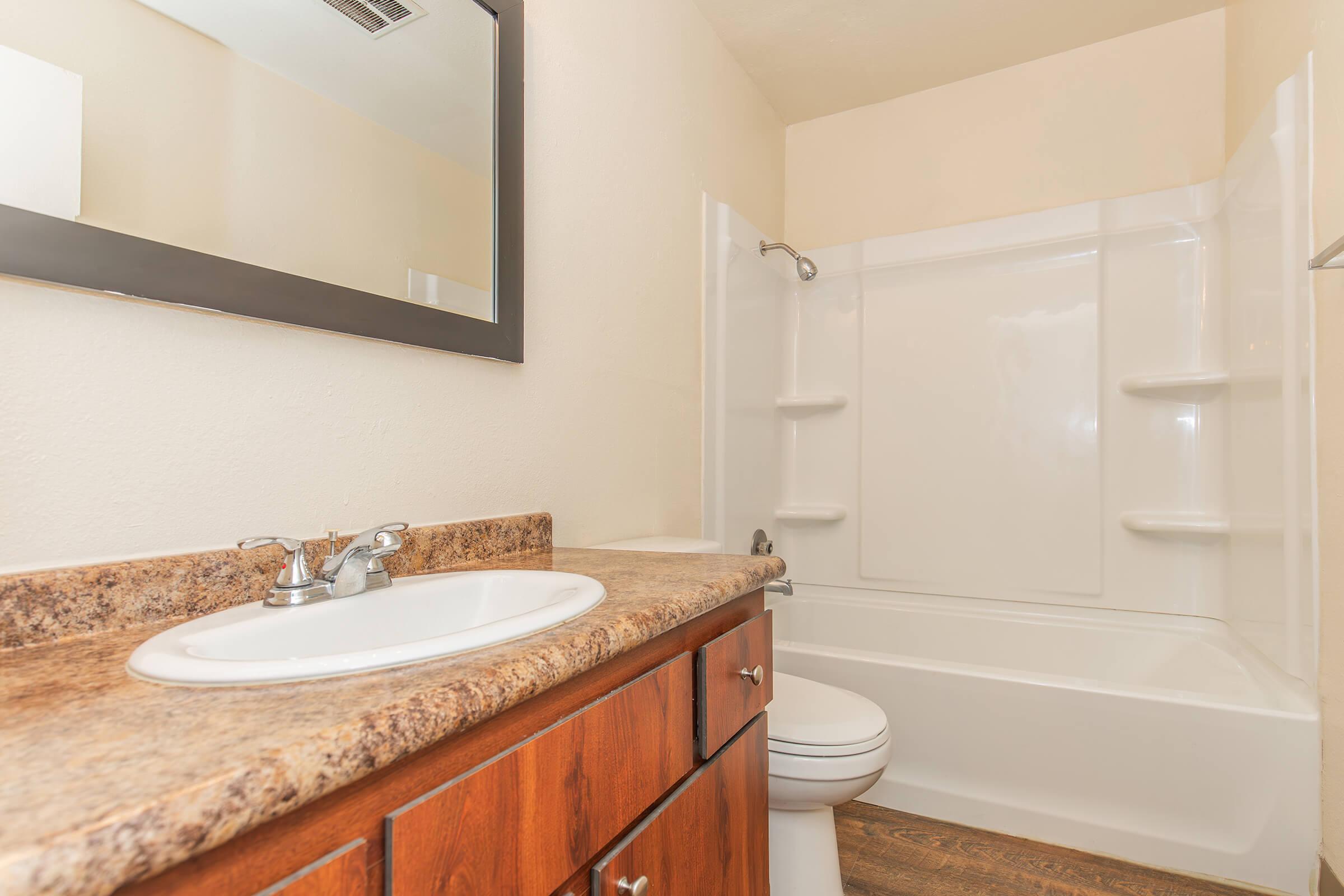 A clean and modern bathroom featuring a white tub and shower combination, a single sink vanity with a granite countertop, a large mirror above the sink, and neutral wall colors. The flooring appears to be a wood-like material, adding warmth to the space.