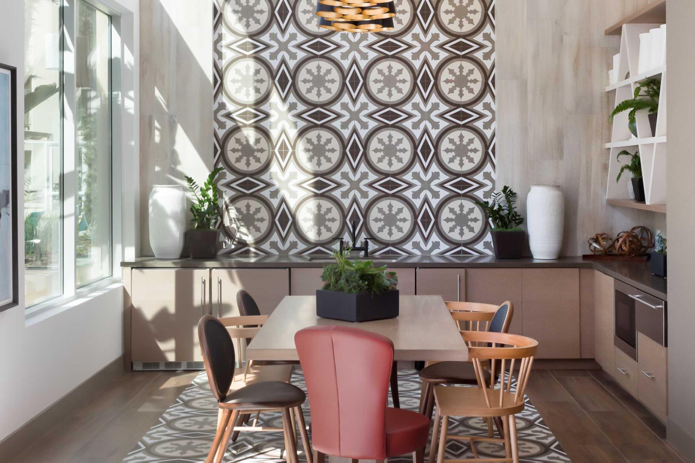 Modern kitchen with a spacious dining area featuring a large table surrounded by wooden chairs, including a red and natural wood chair. The background showcases an intricately patterned wall with decorative elements and potted plants, complemented by ample natural light streaming through large windows.