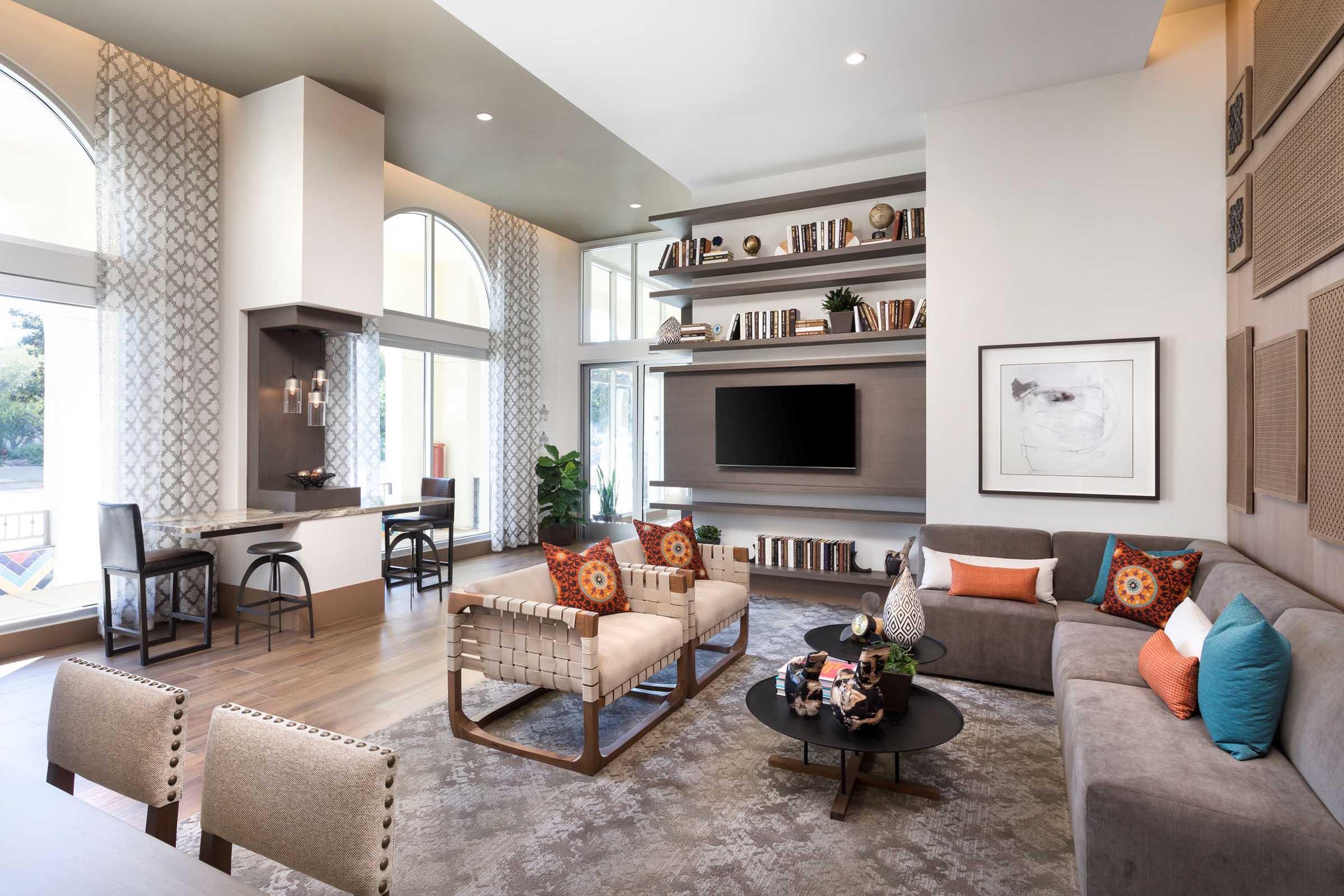 Modern living room featuring a comfortable gray sofa with colorful pillows, a round coffee table with decorative items, and stylish chairs. The walls have a mix of open shelving filled with books and art, large windows letting in natural light, and a cozy dining area visible in the background.