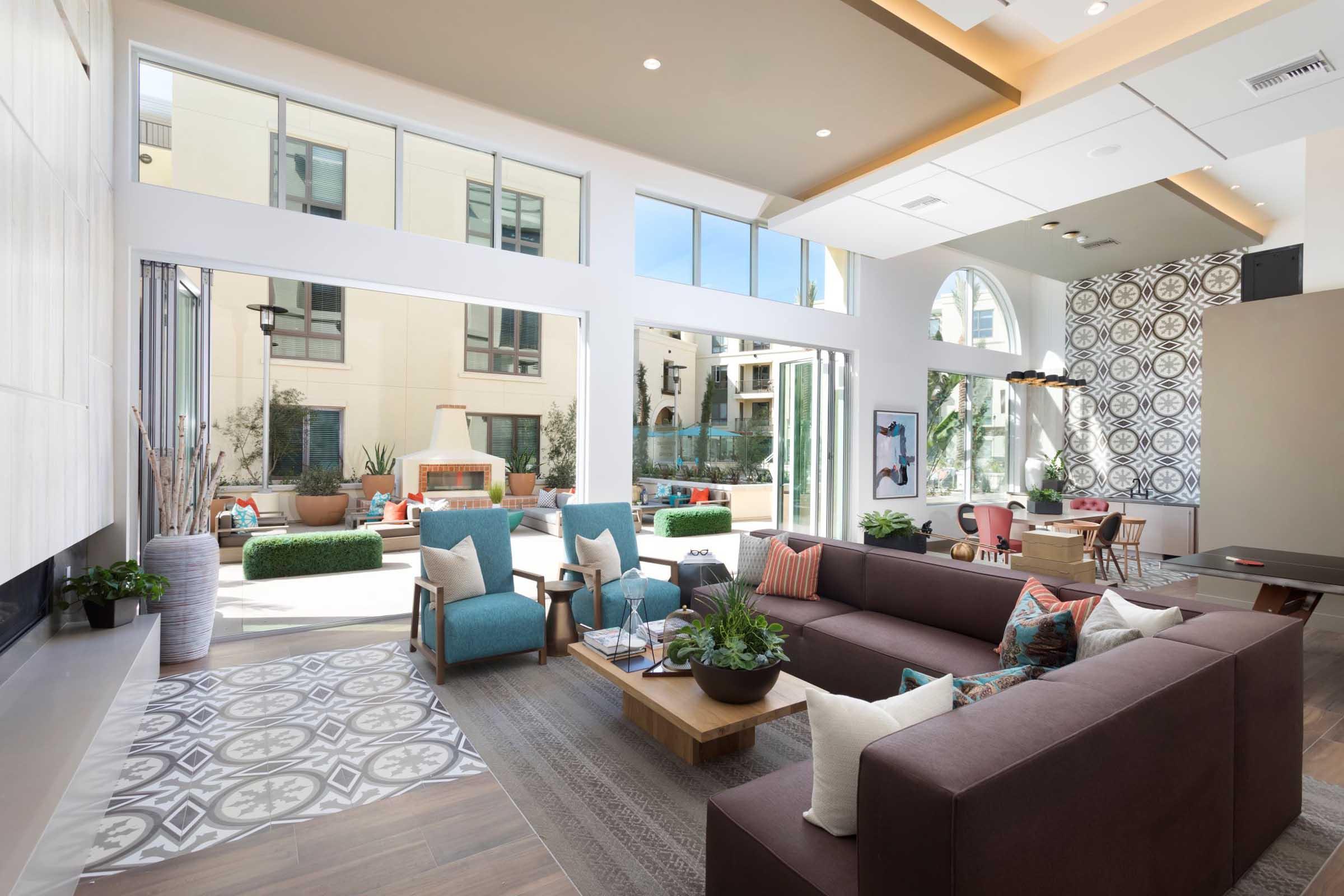 A spacious and modern common area with large windows, featuring a cozy brown sectional sofa, decorative cushions, and a wooden coffee table. The room has stylish decor elements, a fireplace, and a view of an outdoor patio with greenery. Light pours in, creating an inviting atmosphere.