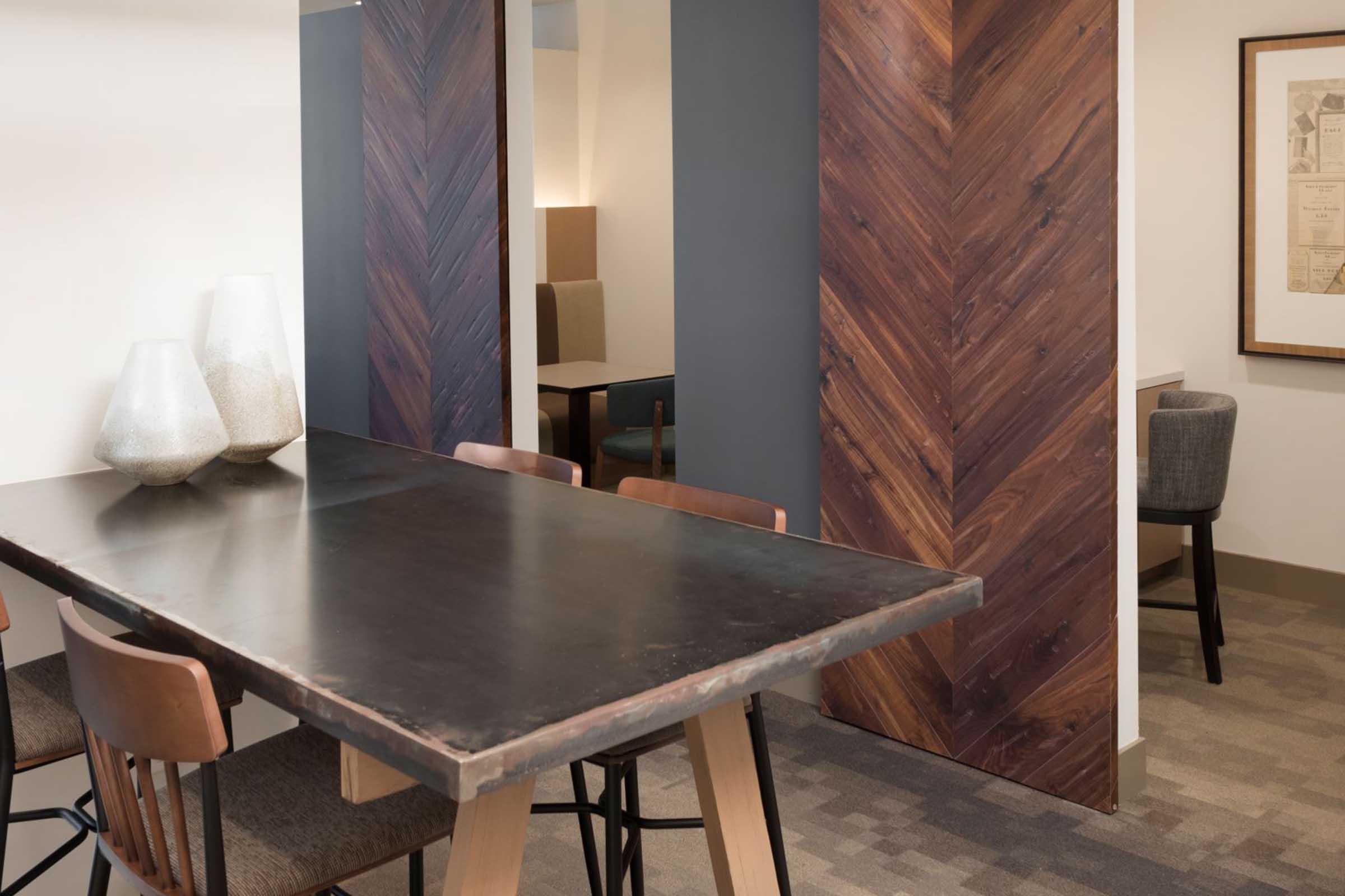 A modern dining space featuring a dark wood table surrounded by brown chairs. In the background, there are decorative vases and a textured wooden wall with a herringbone pattern. Soft lighting creates a warm atmosphere, and a framed artwork is partially visible on the wall.