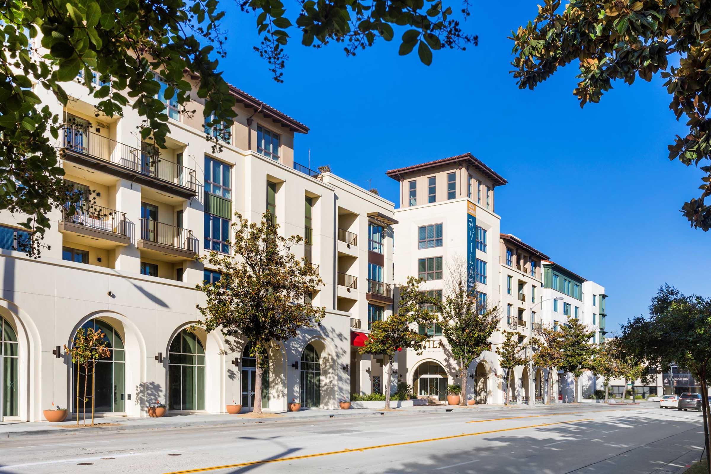Modern multi-story buildings with balconies and large windows line a well-maintained street. Trees with green foliage are visible along the sidewalks, and the clear blue sky provides a bright backdrop, creating a welcoming urban scene.