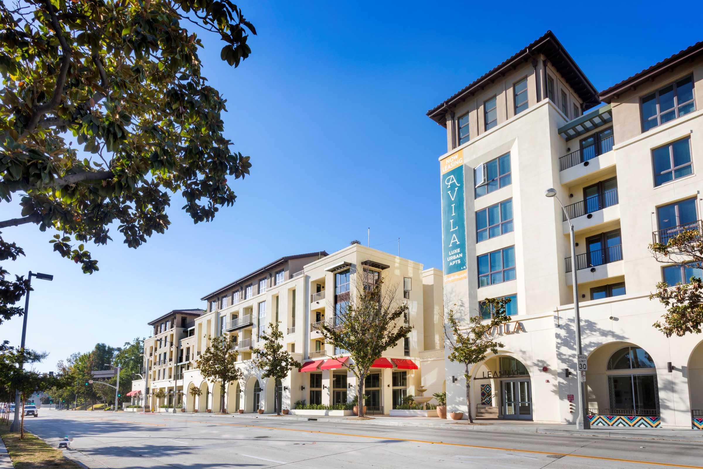 A modern multi-story building with a cream-colored facade, large windows, and balconies. The ground level features red awnings, outdoor seating, and storefronts. A sign reading "Avila" is prominently displayed. The surrounding area includes trees and a clear blue sky.