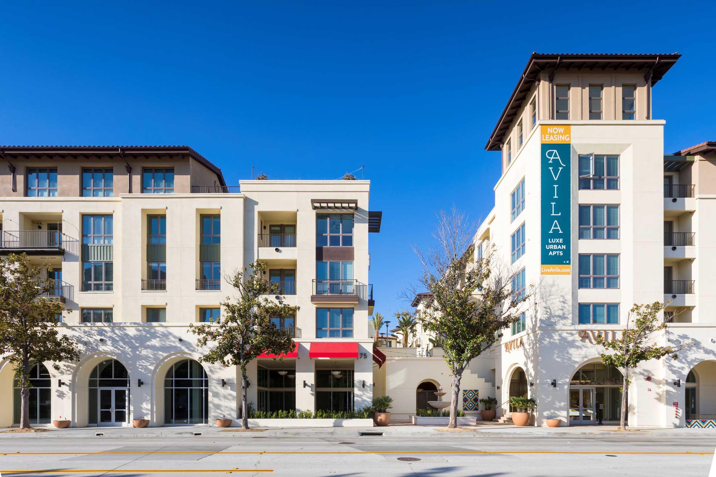 Modern multi-story apartment buildings with a mix of architectural styles, featuring large windows and balconies. A bright blue sky is in the background. A prominent banner advertising "NOW LEASING AVILA LUXE URBAN APTS" hangs from one of the buildings. Trees line the street in front.