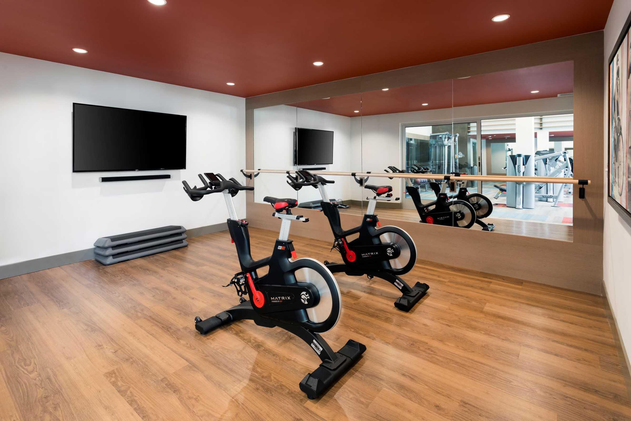 A modern fitness room featuring two stationary bikes, a large wall-mounted TV, and mirrors. The space has wood flooring and an accent wall painted in a warm color, creating an inviting atmosphere for exercise. Additional gym equipment can be seen in the background.