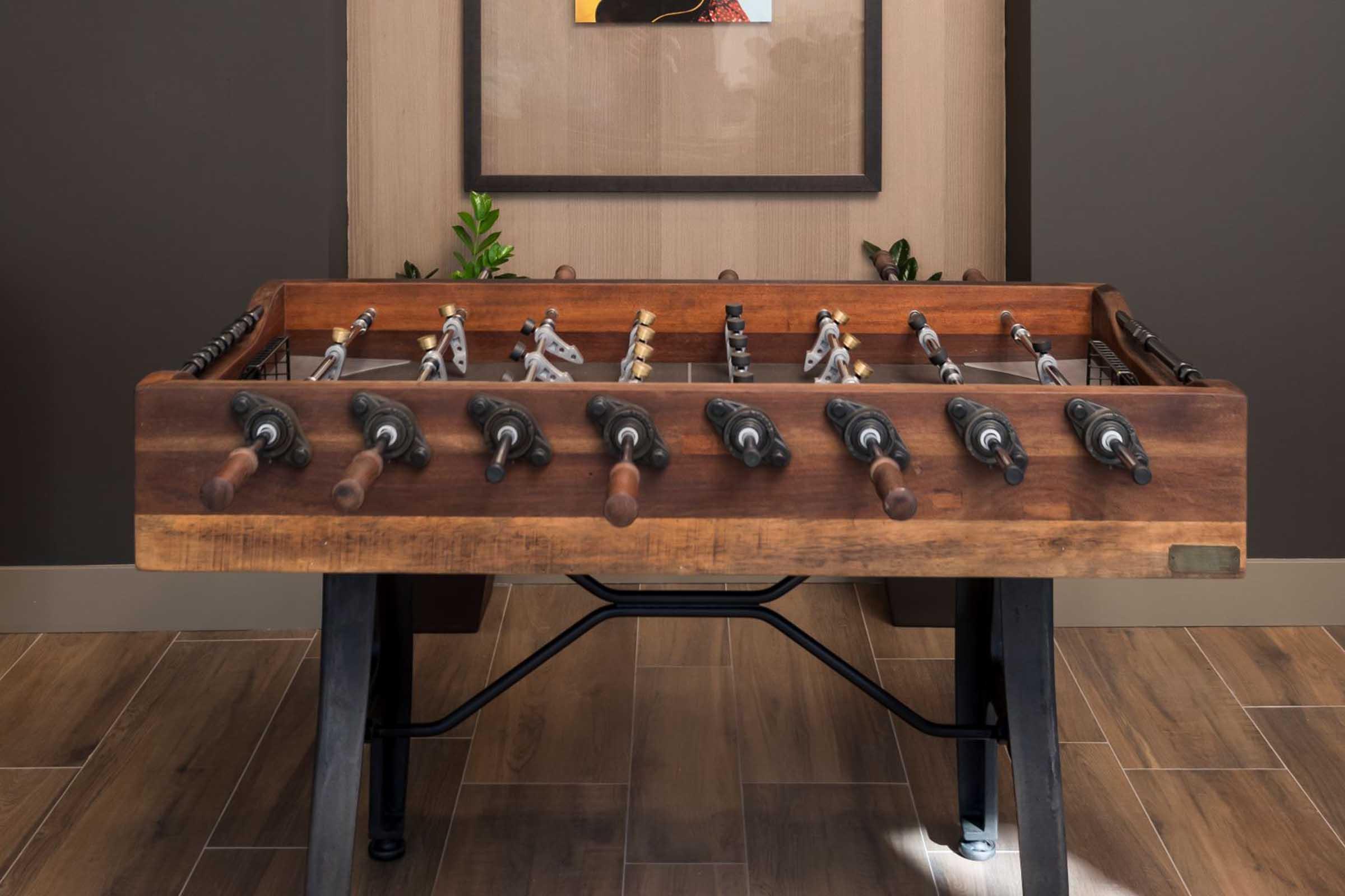 A wooden foosball table with black and white players, situated in a modern room with dark walls and a plant in the background. A piece of artwork is visible on the wall above the table, adding a decorative touch to the space. The floor is made of large, light-colored tiles.