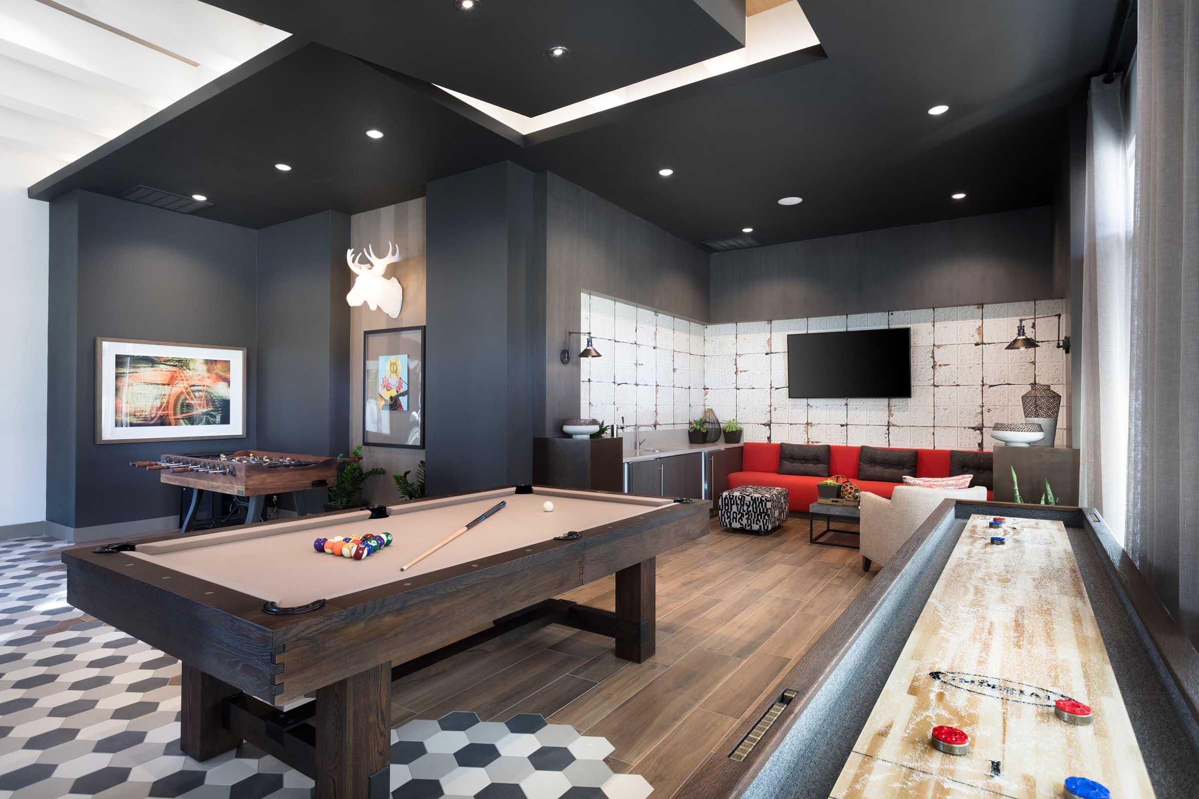 A stylish game room featuring a pool table, foosball table, and an air hockey table. The room has modern decor with gray and red accents, large windows, and artistic wall art, creating a cozy yet vibrant atmosphere for recreational activities.