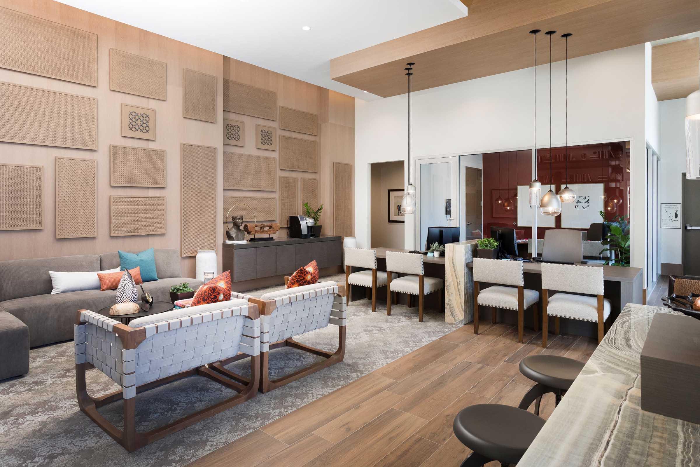 A modern, stylish living space featuring a cozy gray sofa with colorful cushions, unique wall art, and sleek wood accents. The room includes a contemporary dining area with white chairs and decorative lights, and a small kitchen area in the background, all contributing to a warm and inviting atmosphere.